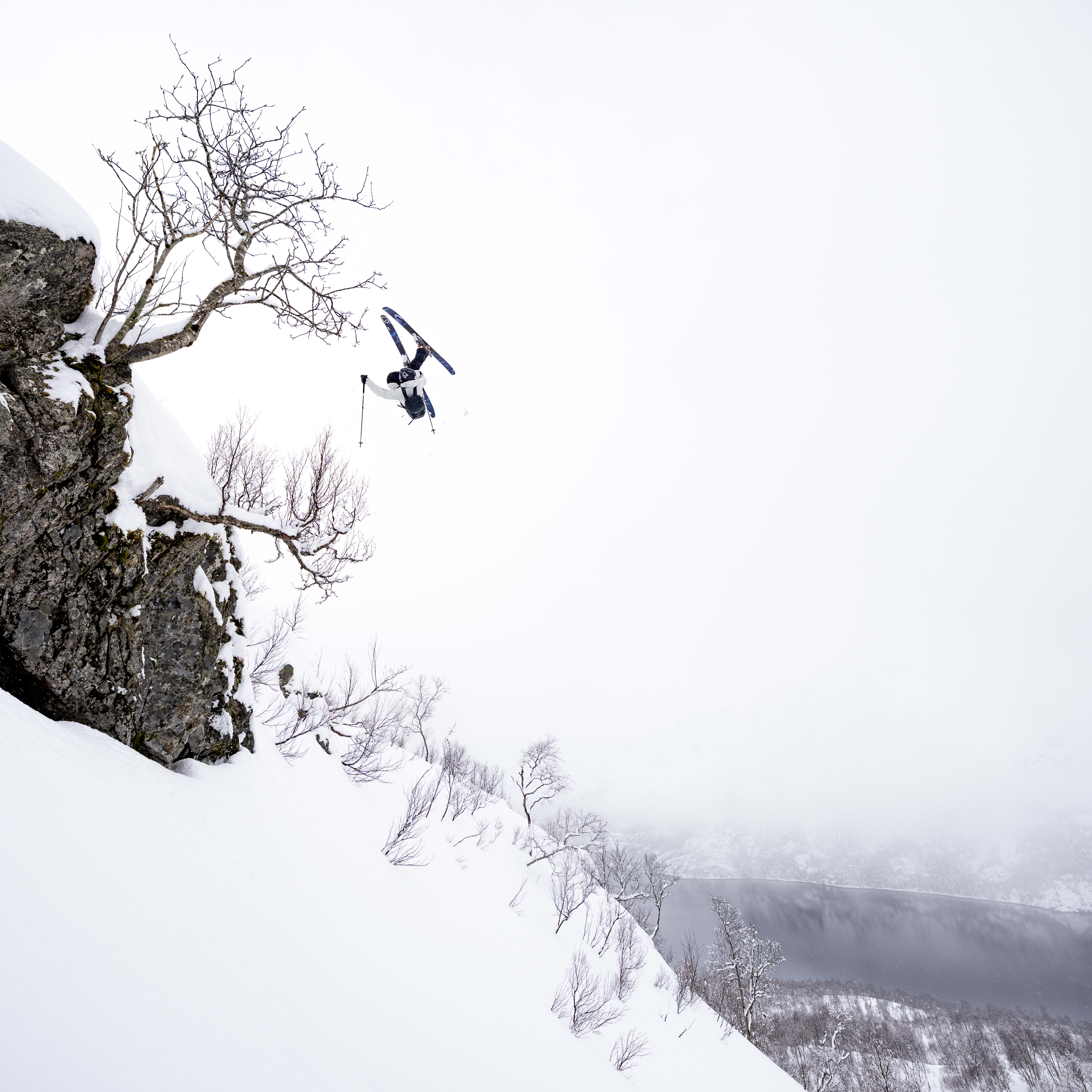 A skier wearing Norrøna clothing performs a backflip off a cliff, with a snowy mountain range and a lake in the background.
