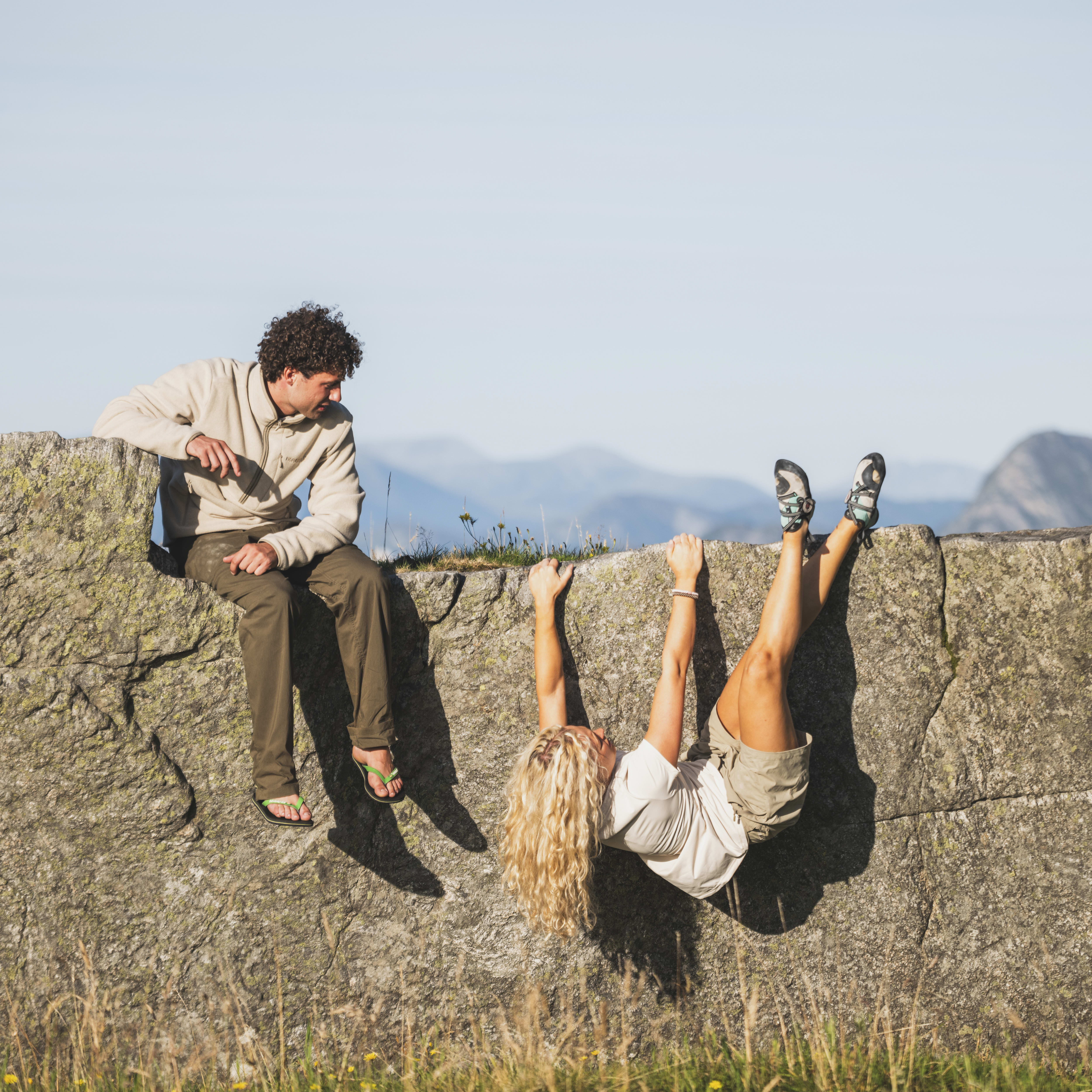 A person in a Norrøna fleece jacket sits on a rock wall looking down at another person hanging upside down. Mountains are in the background.