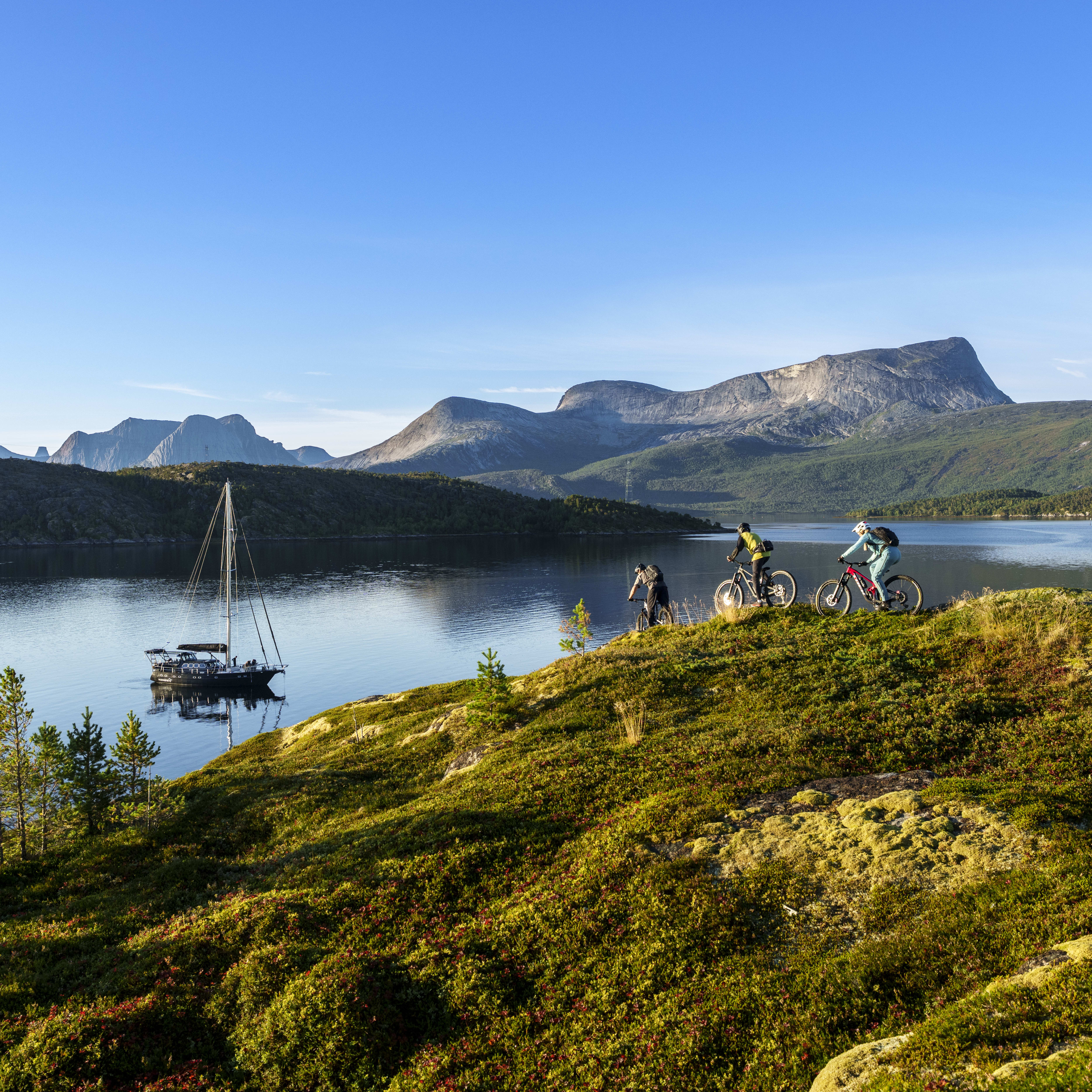 Three mountain bikers, one wearing Norrøna clothing, ride along a grassy ridge overlooking a lake with a sailboat. Mountains in the background.