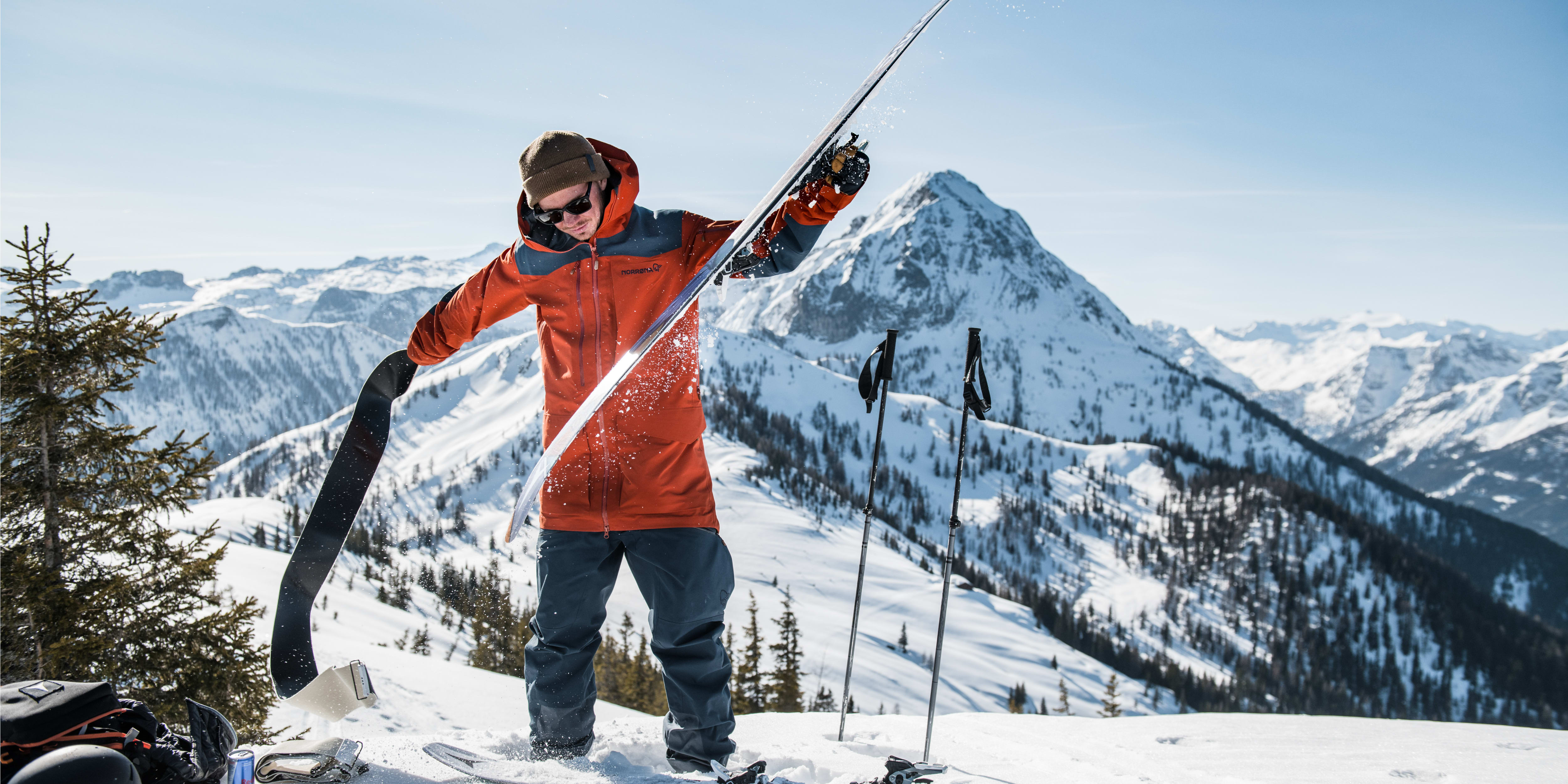 A man wearing a Norrøna jacket stands on a snowy mountaintop with his skis and poles. 
