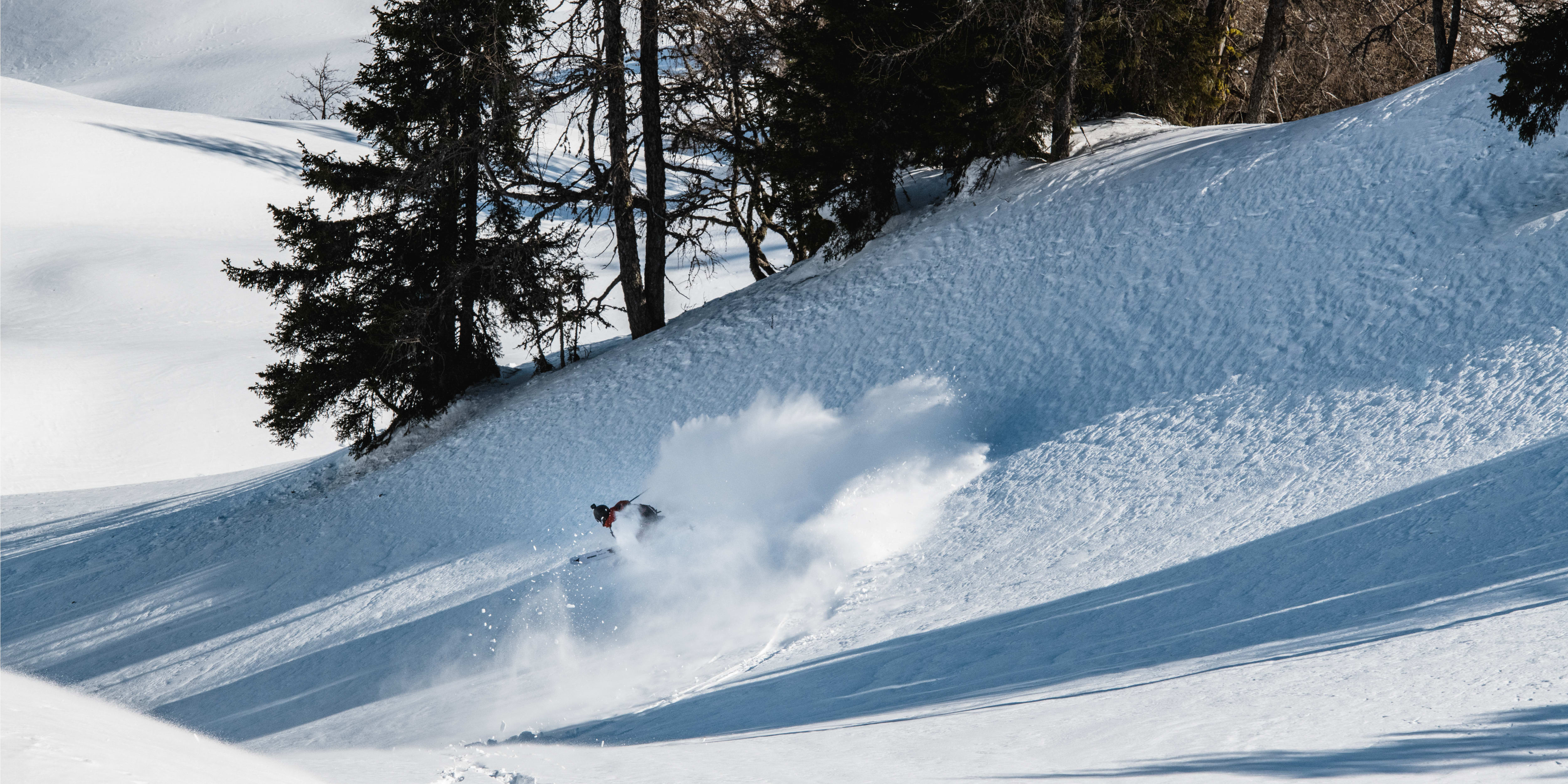 A person wearing a Norrøna ski suit is skiing down a snowy slope, kicking up snow.  
