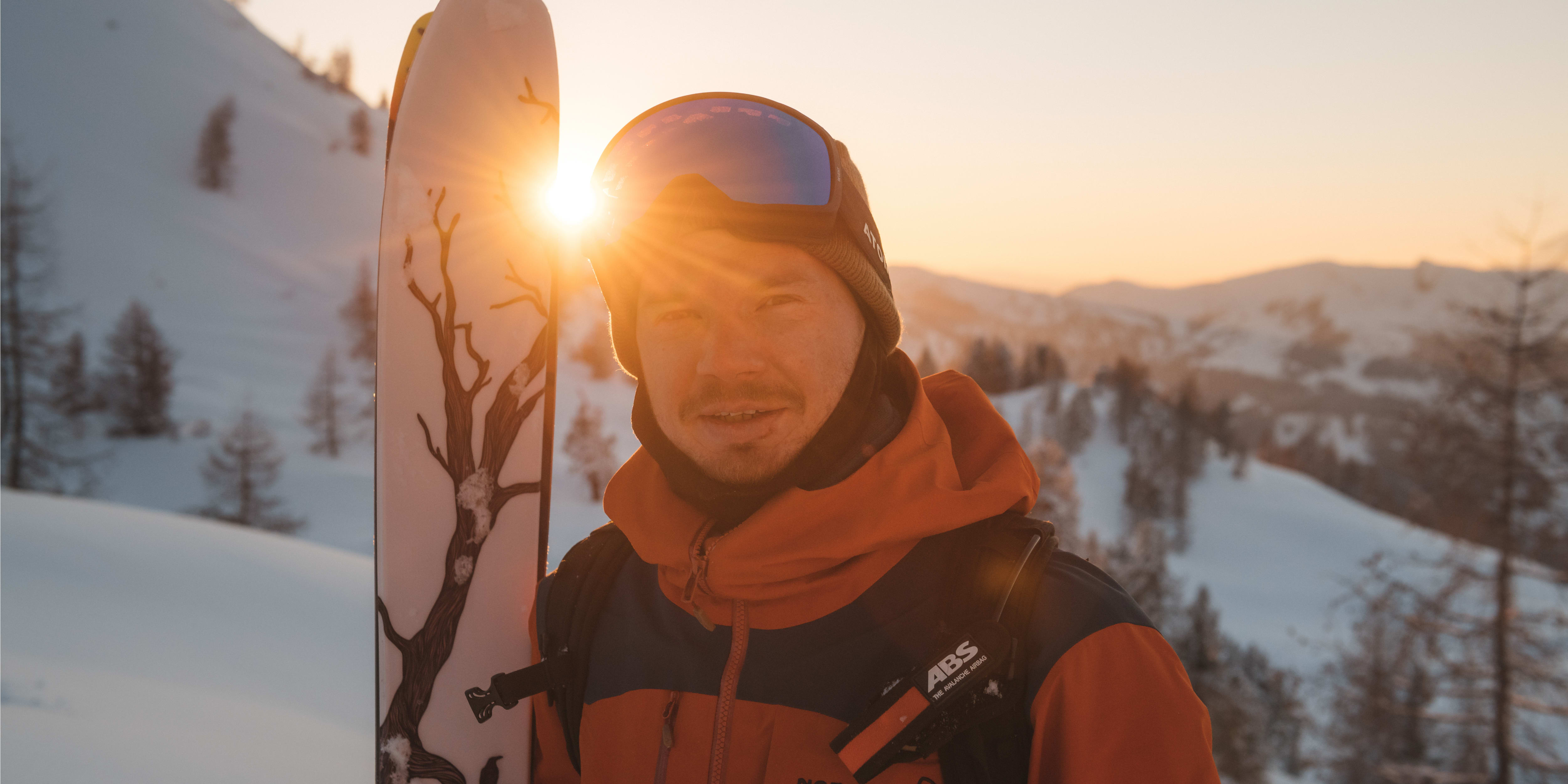 A man wearing a Norrøna jacket and a ski helmet stands in snowy mountains. The sun shines brightly behind him, creating a warm glow. He is holding a ski. 
