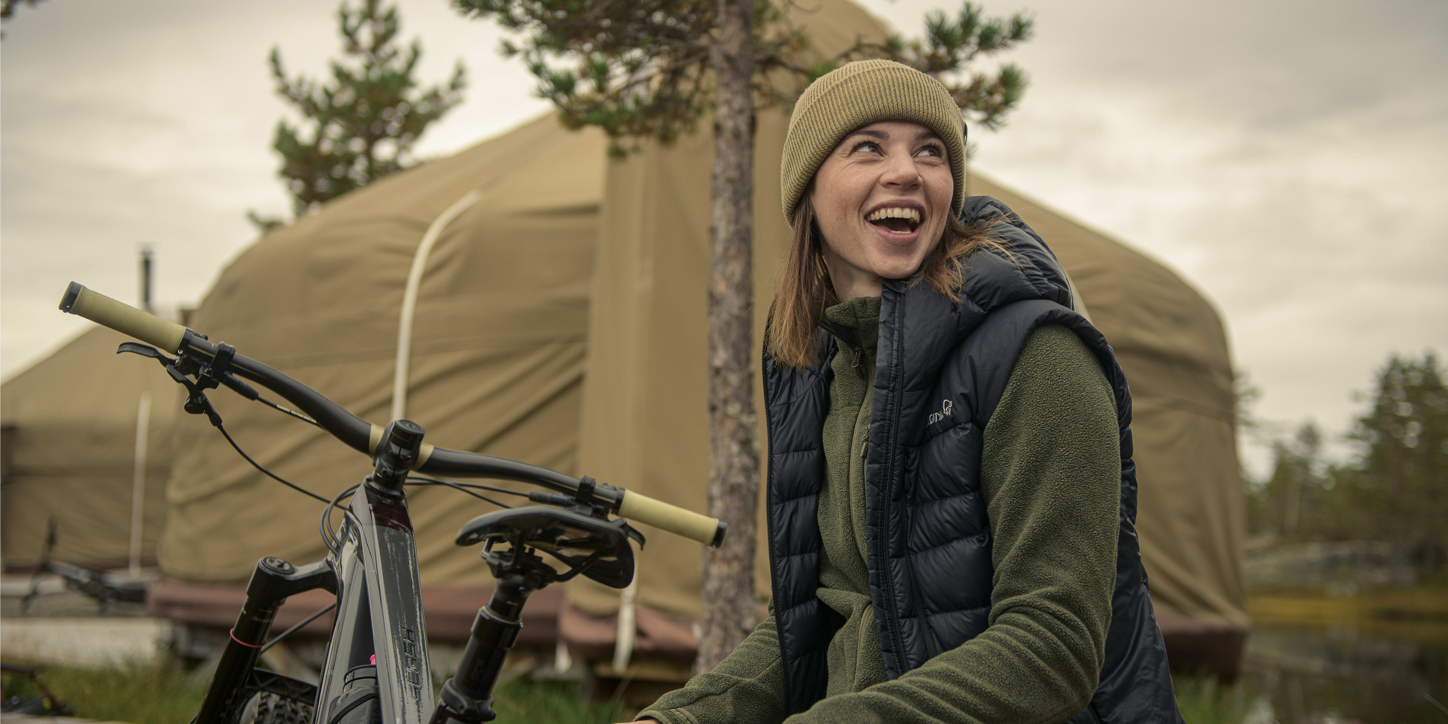 A woman smiles, wearing a Norrøna vest and beanie, next to her mountain bike. A large canvas tent and trees are in the background.
