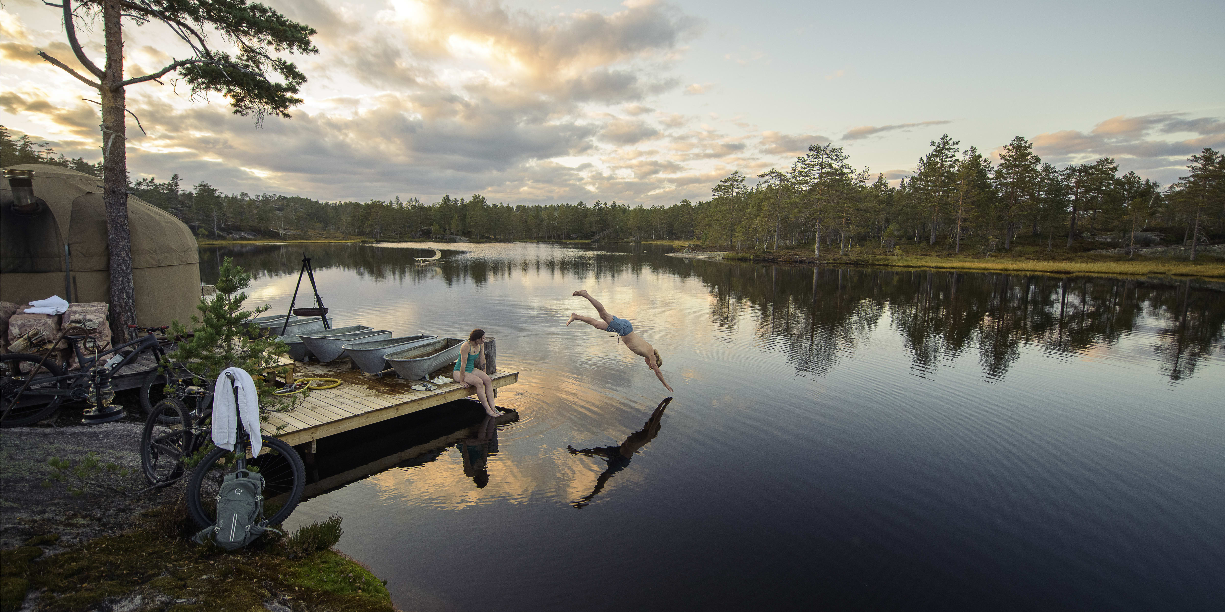 Person jumps into a lake from a wooden dock. A person sits on the dock.  A Norrøna backpack is on the ground beside a bike.  

