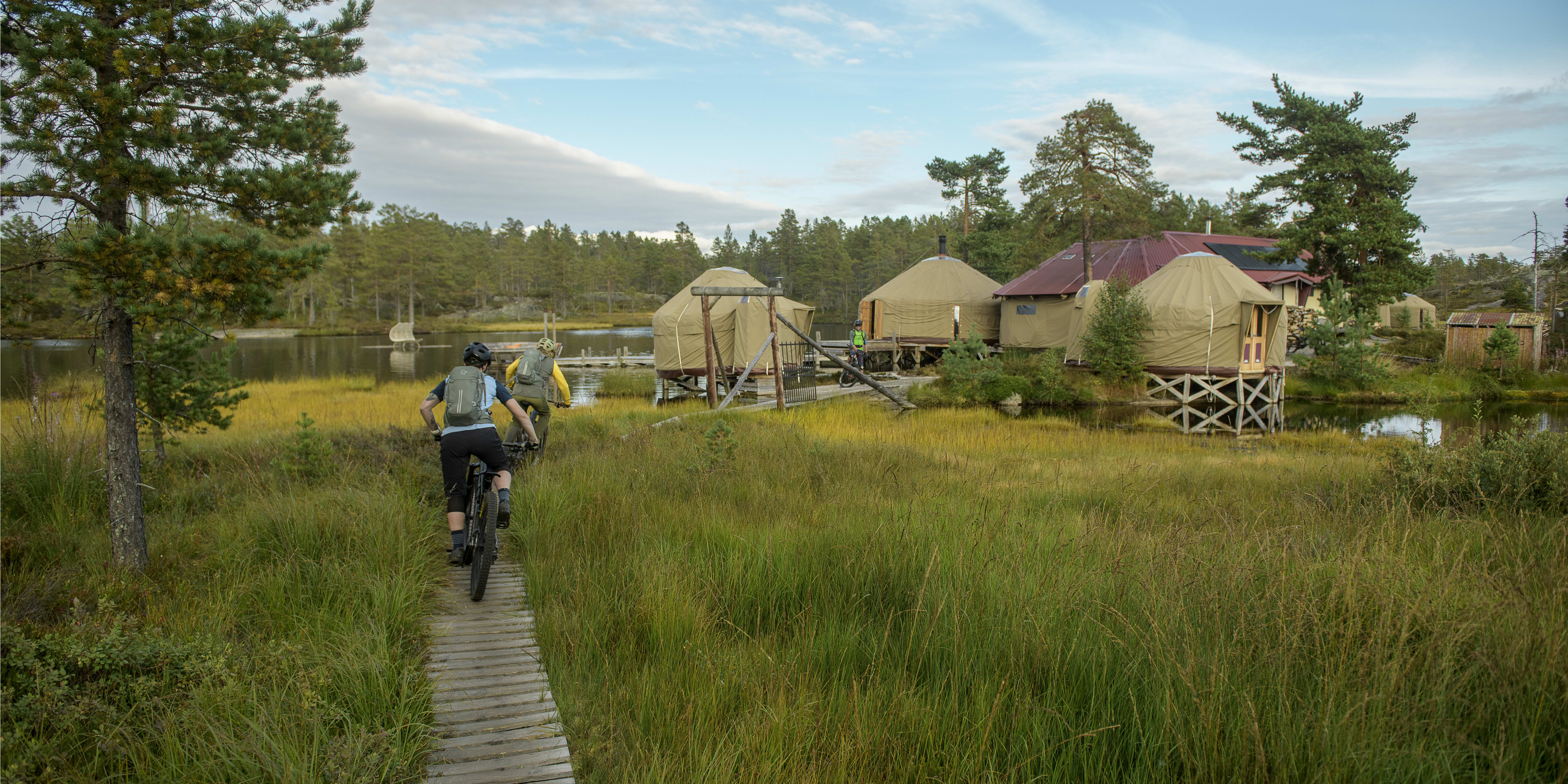 Two people riding mountain bikes on a wooden path in a grassy field. They are passing by a lake and some cabins. One biker is wearing a Norrøna backpack. 
