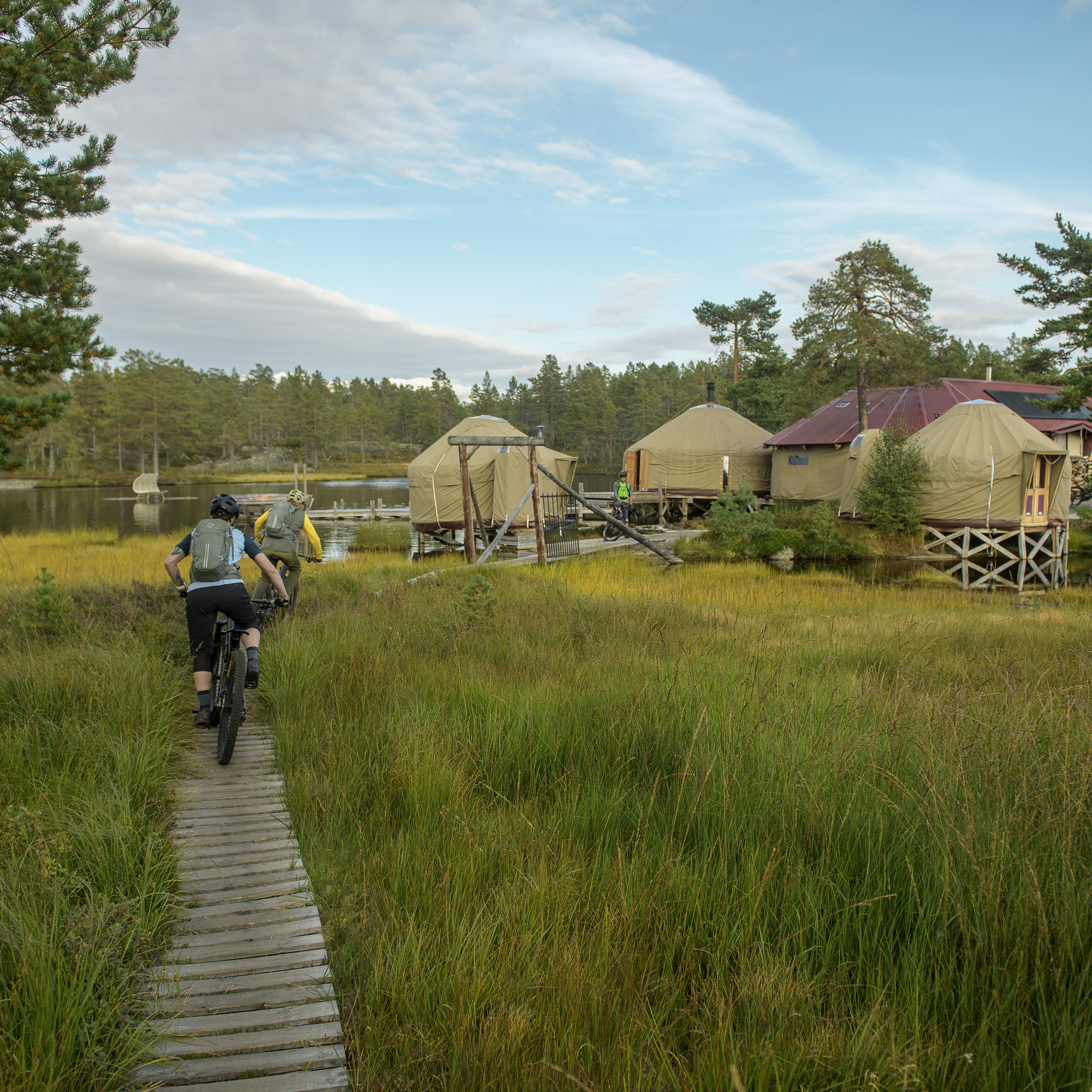 Two people riding mountain bikes on a wooden path in a grassy field. They are passing by a lake and some cabins. One biker is wearing a Norrøna backpack. 
