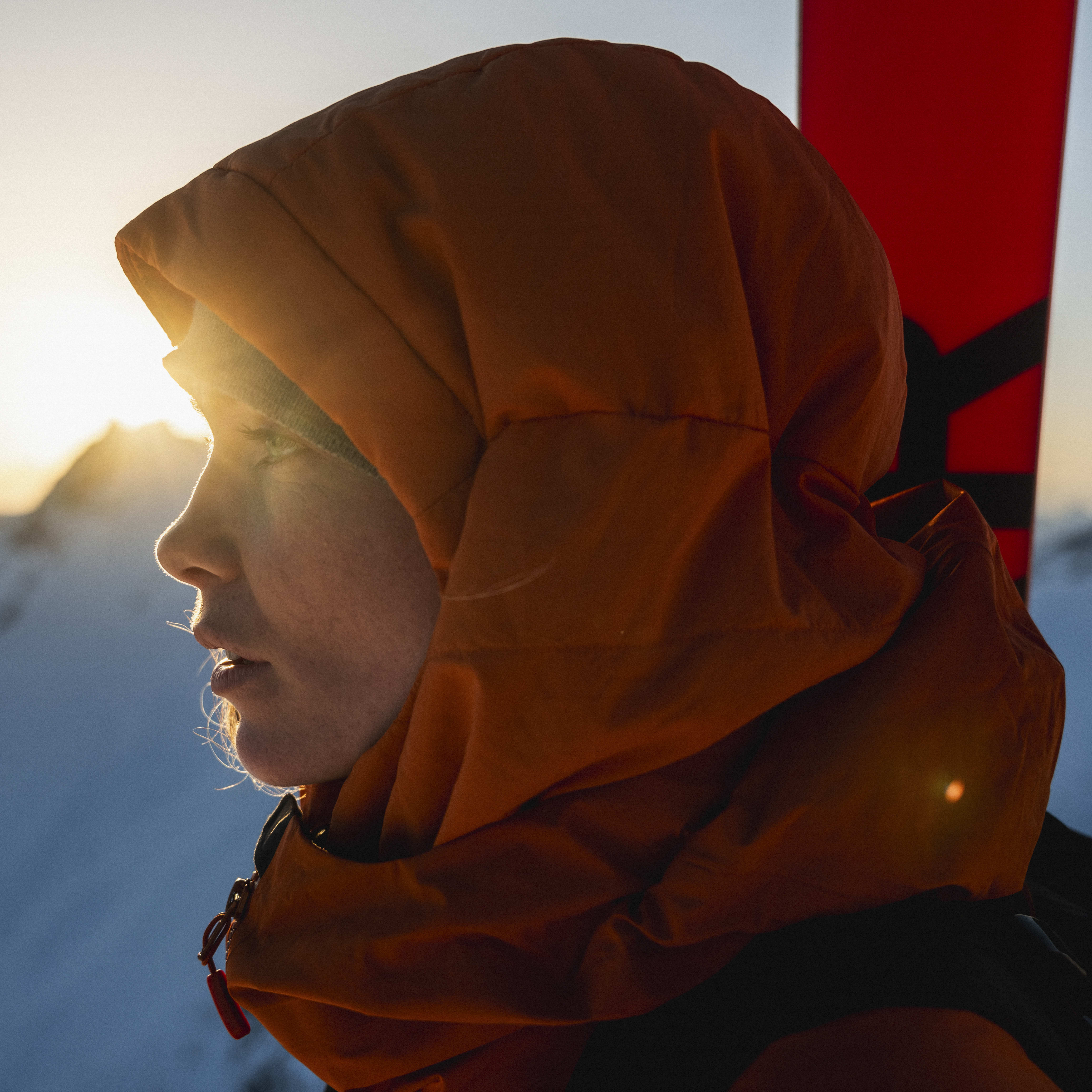 Profile of a person in a Norrøna hooded jacket with skis against a snowy mountain backdrop at sunrise.