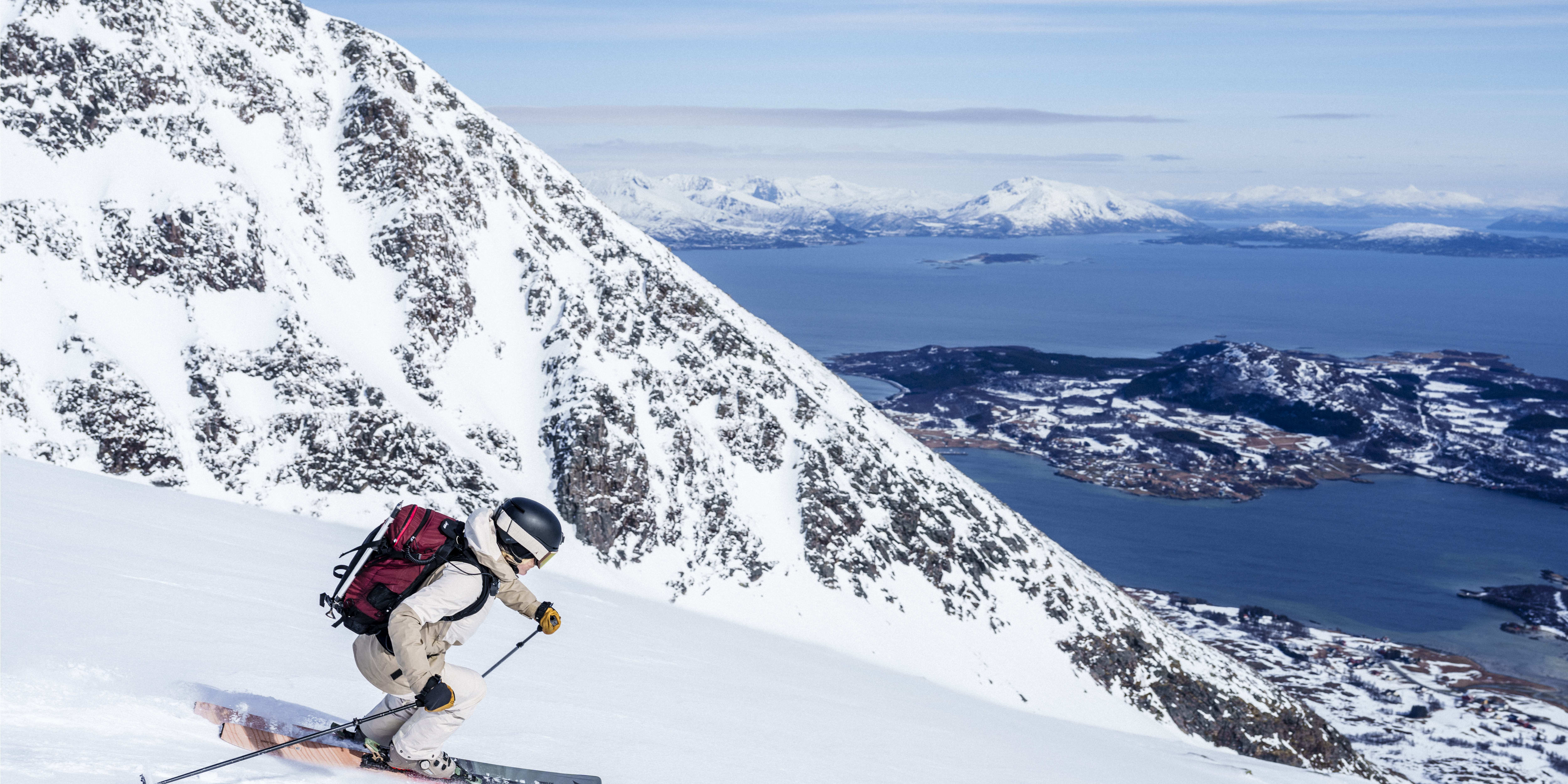 A skier wearing a red backpack and helmet skis down a snow-covered mountainside. The skier is in the foreground with a blue ocean and snow-covered mountains in the background.
