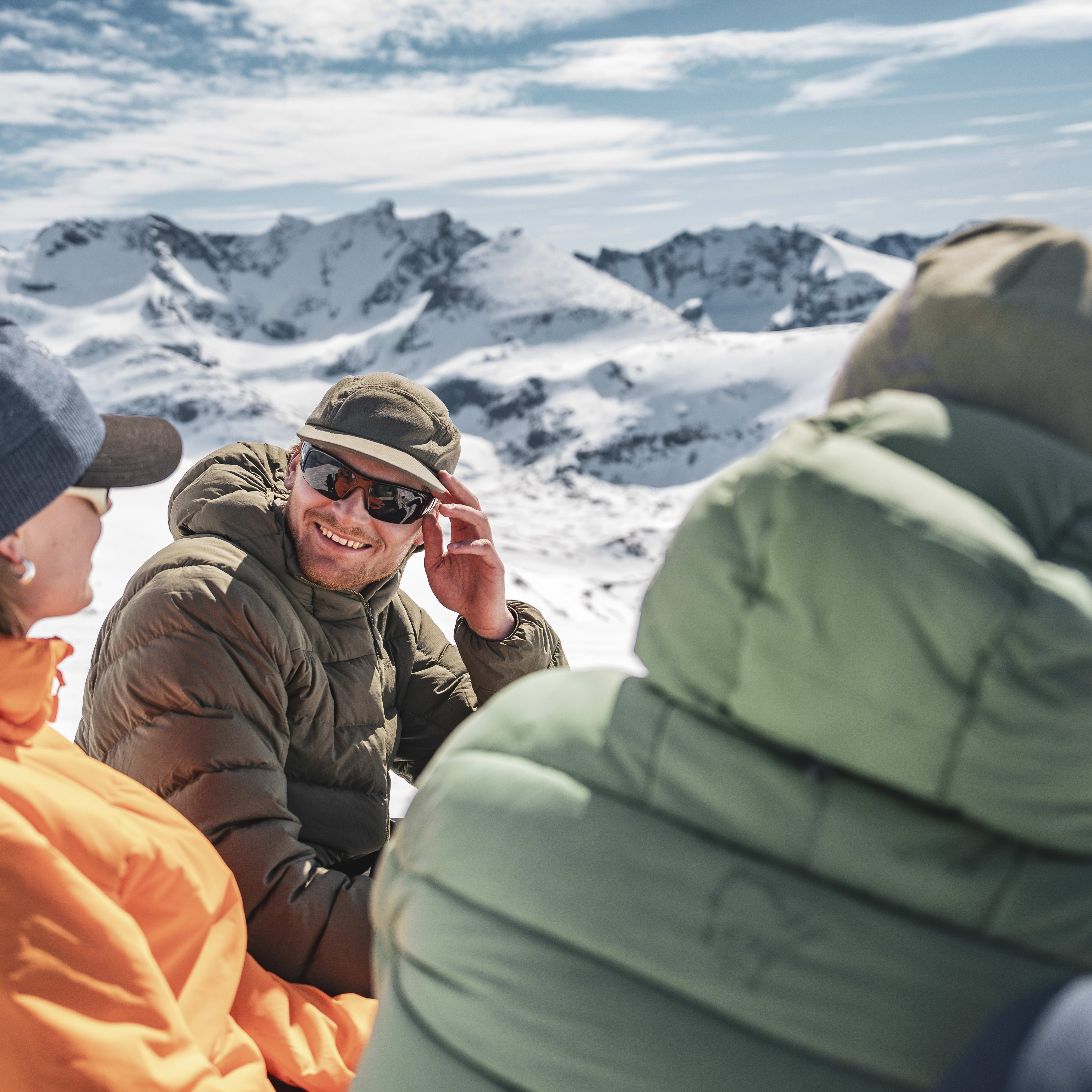 Three people in winter clothing sitting on a snowy mountain. The person in the middle is wearing sunglasses and a Norrøna jacket.  
