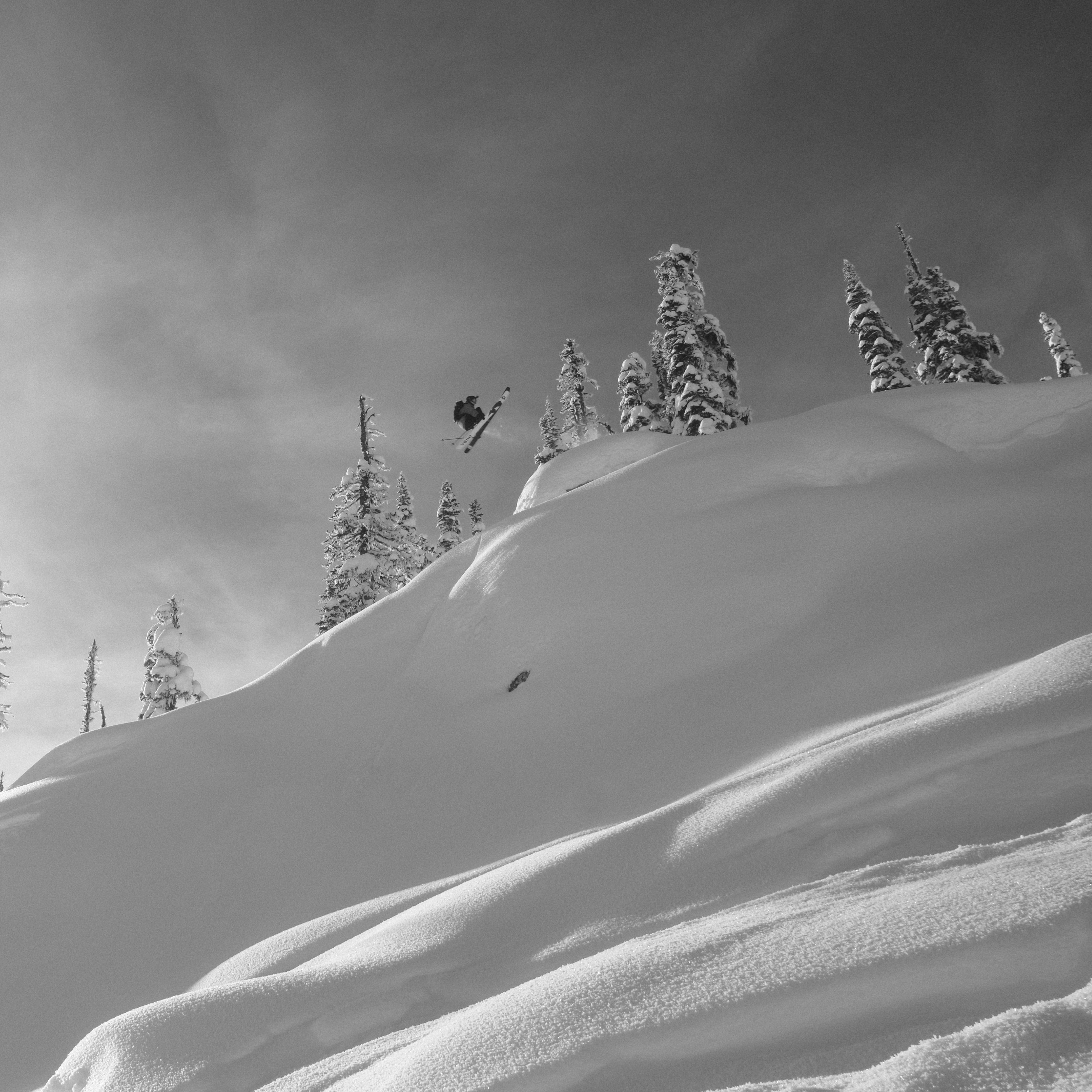A black and white shot of a skier in Norrøna gear mid-air, soaring over a snowy mountain landscape with scattered trees.