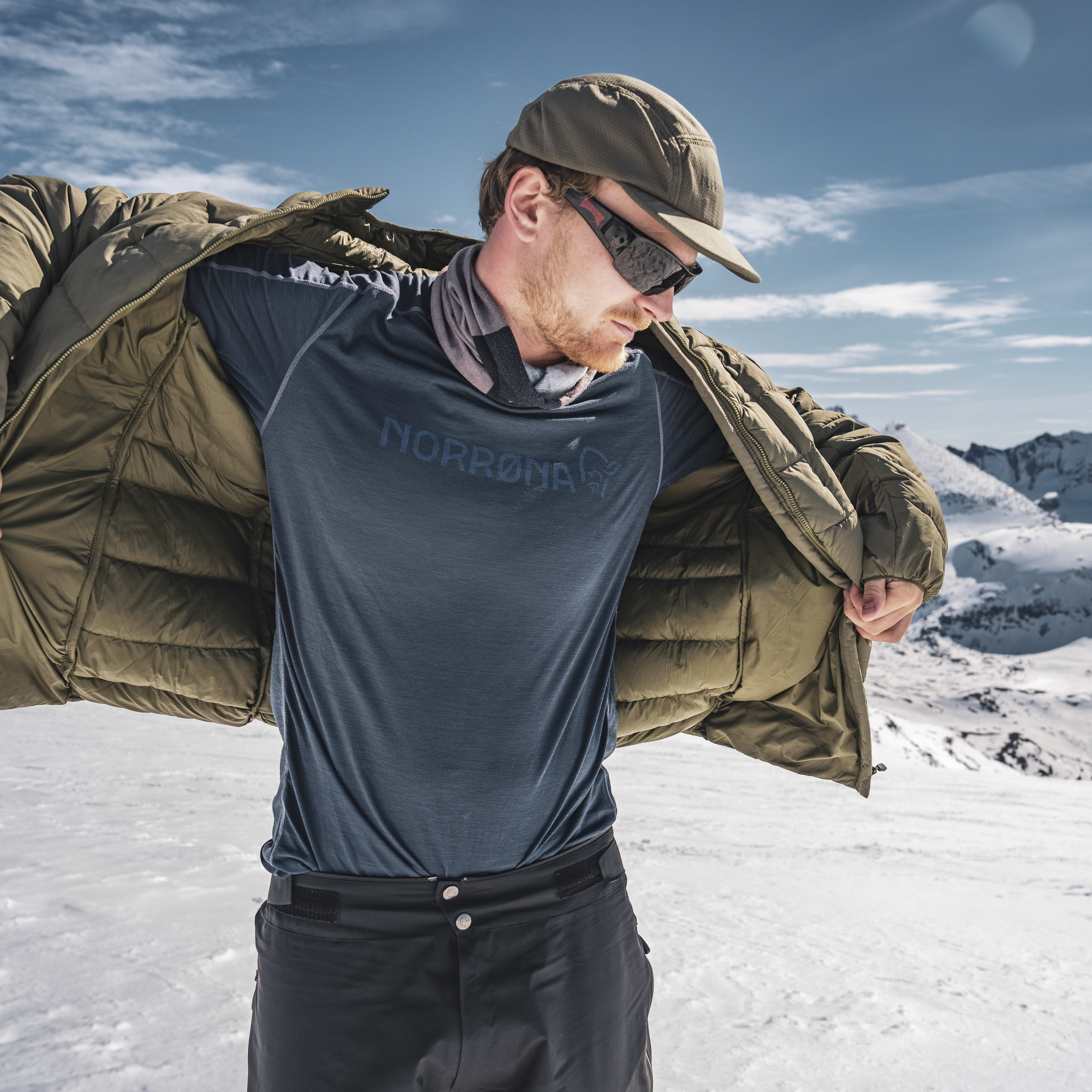 Man in a Norrøna jacket and black pants stands in snowy mountains with a blue sky in the background.