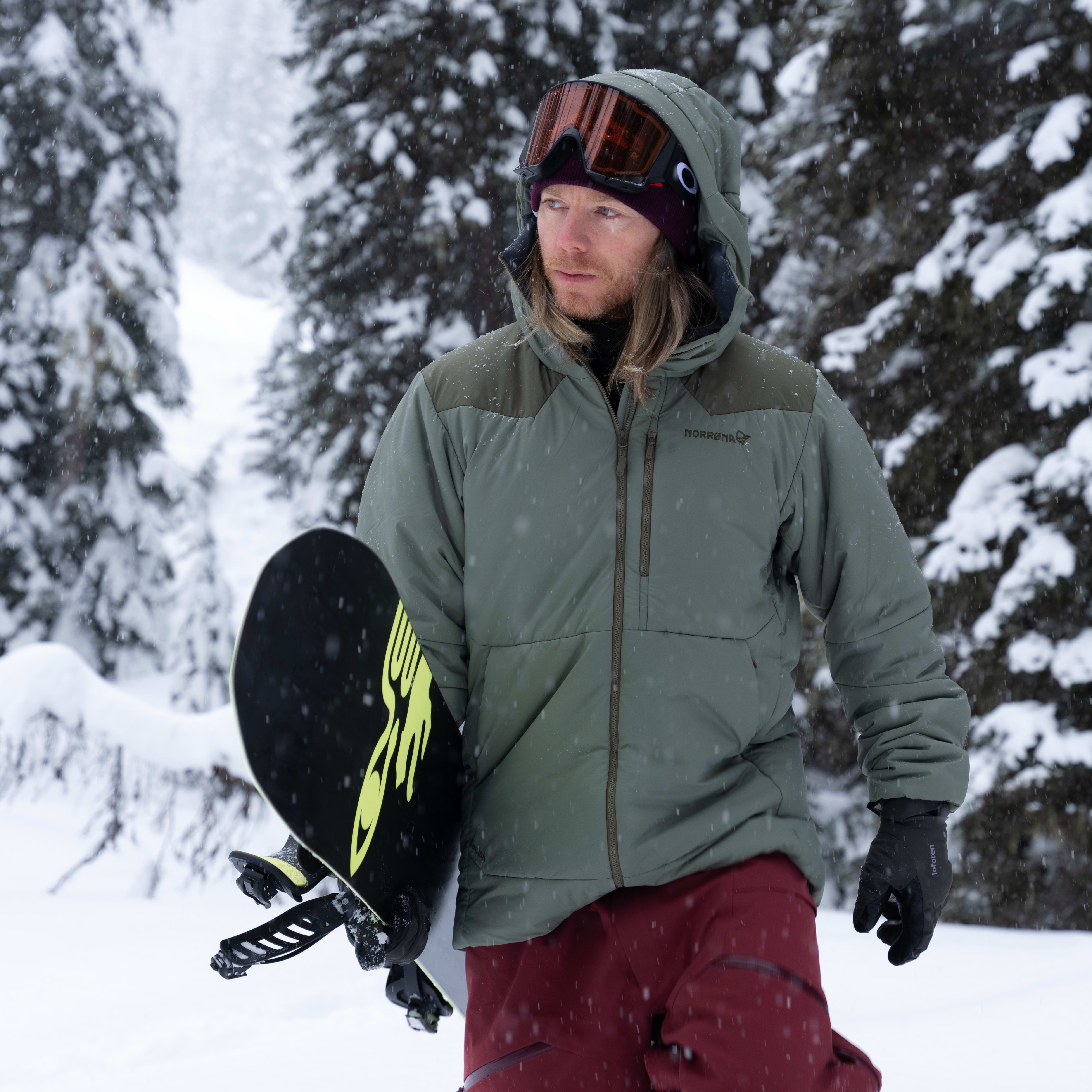 A snowboarder walks through a snowy forest wearing a Norrøna jacket and goggles, carrying their board.