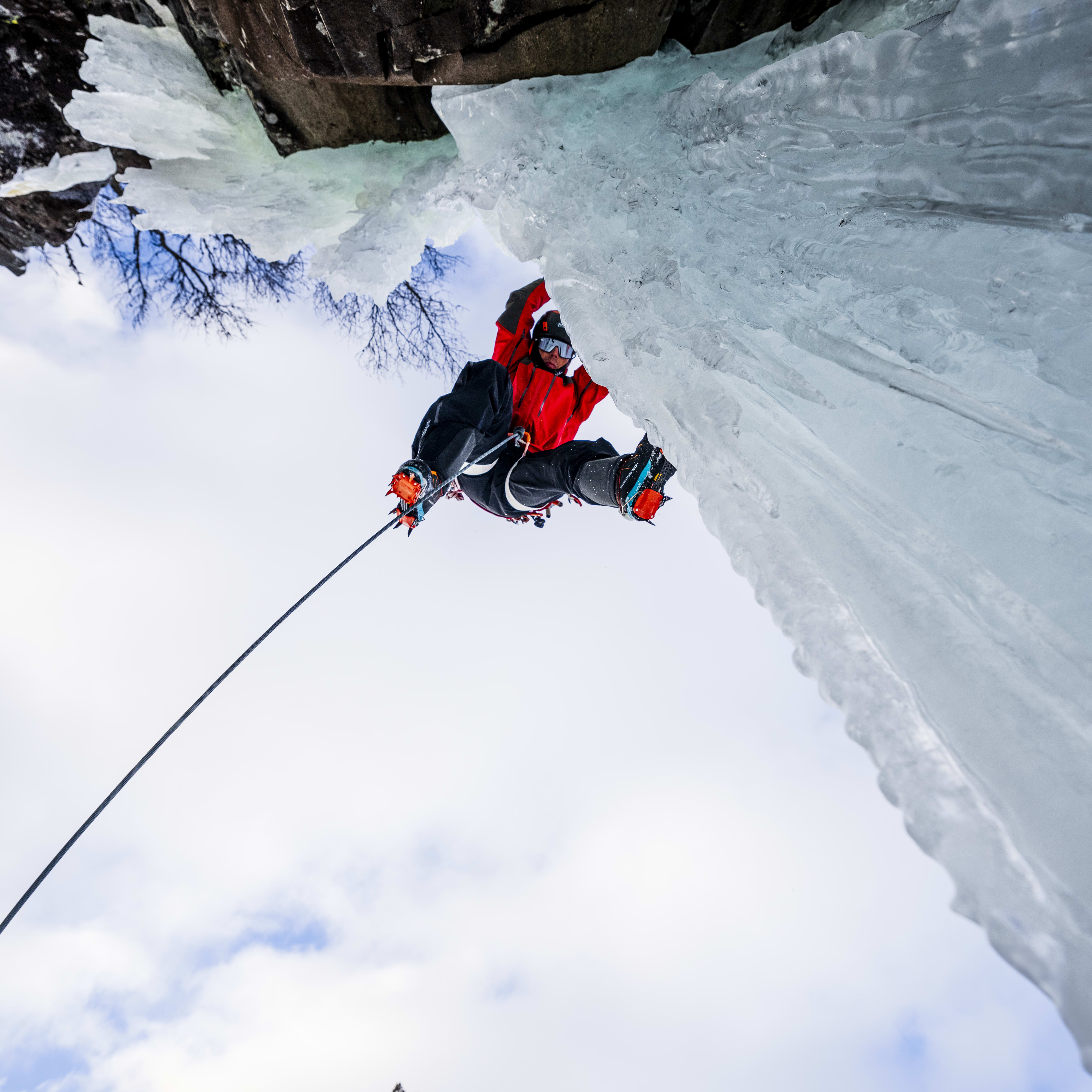 A climber in red Norrøna gear hangs from a rope while ice climbing. 
