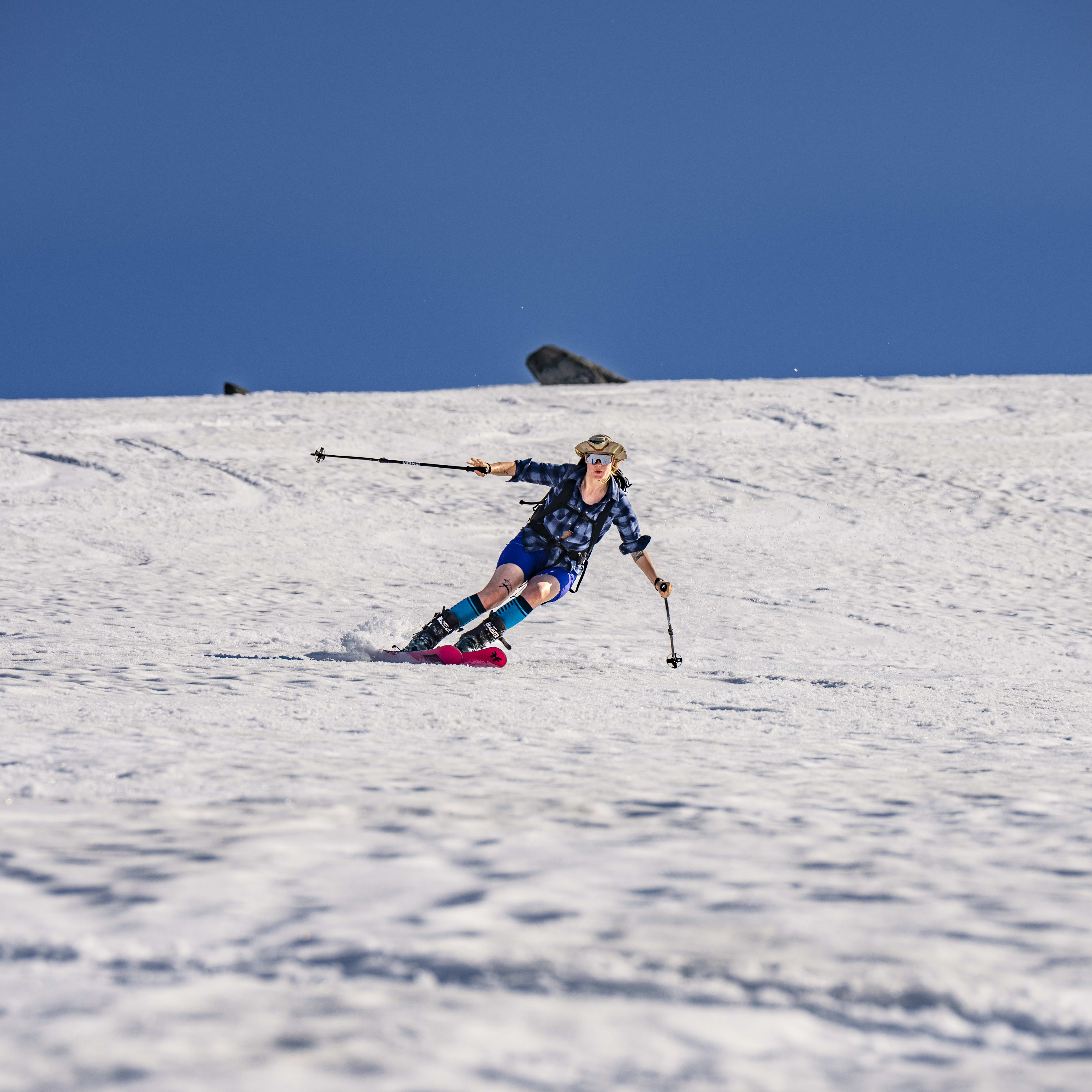 A person wearing a blue plaid shirt and pink ski boots is skiing downhill in a snowy area.  They are holding ski poles,  wearing sunglasses and a hat, and leaving a trail of snow behind them.
