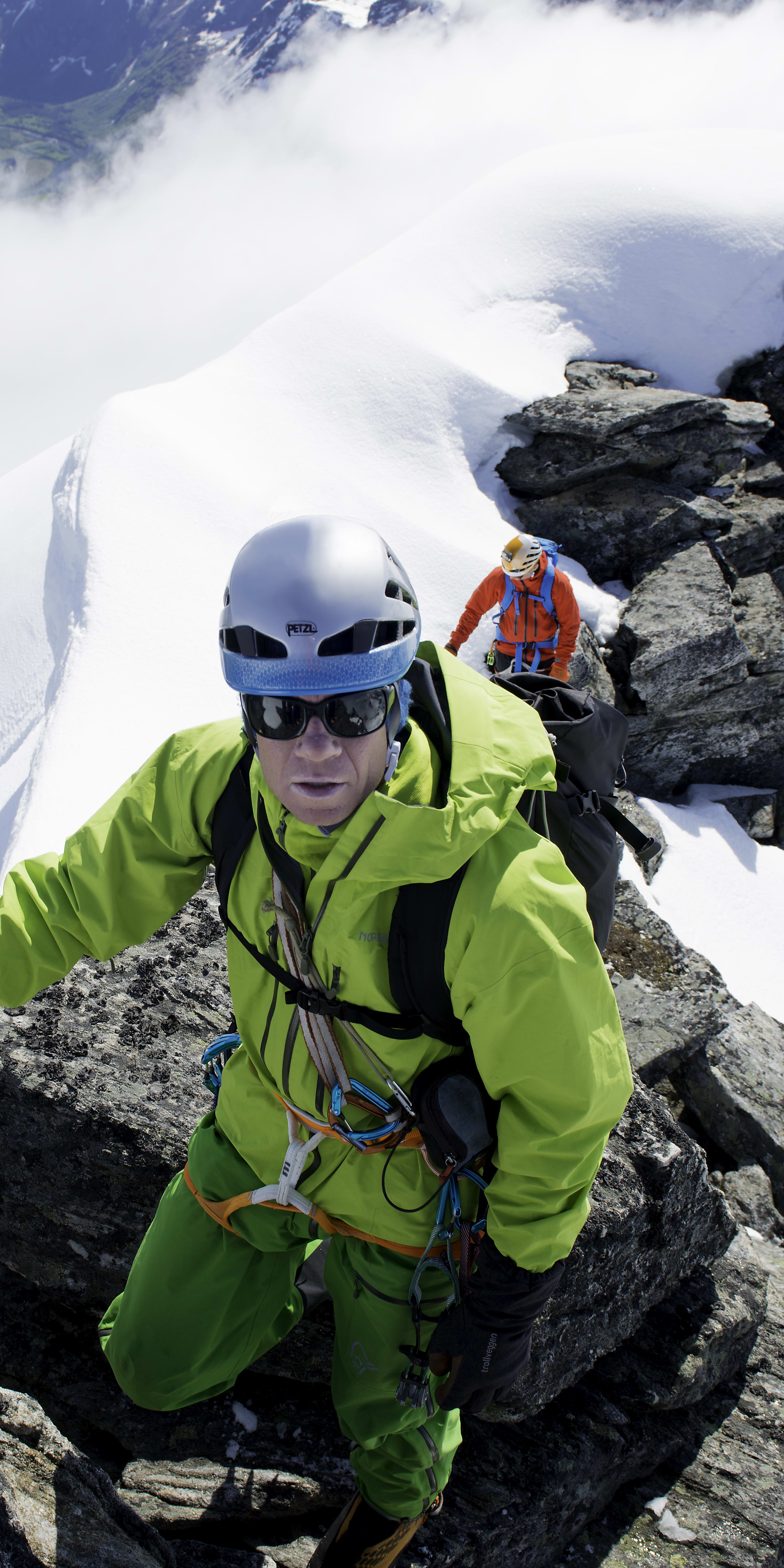 A climber wearing a Norrøna jacket and helmet is on a snowy mountain peak. He's holding onto an ice axe.  The view behind him is a snowy valley. 
