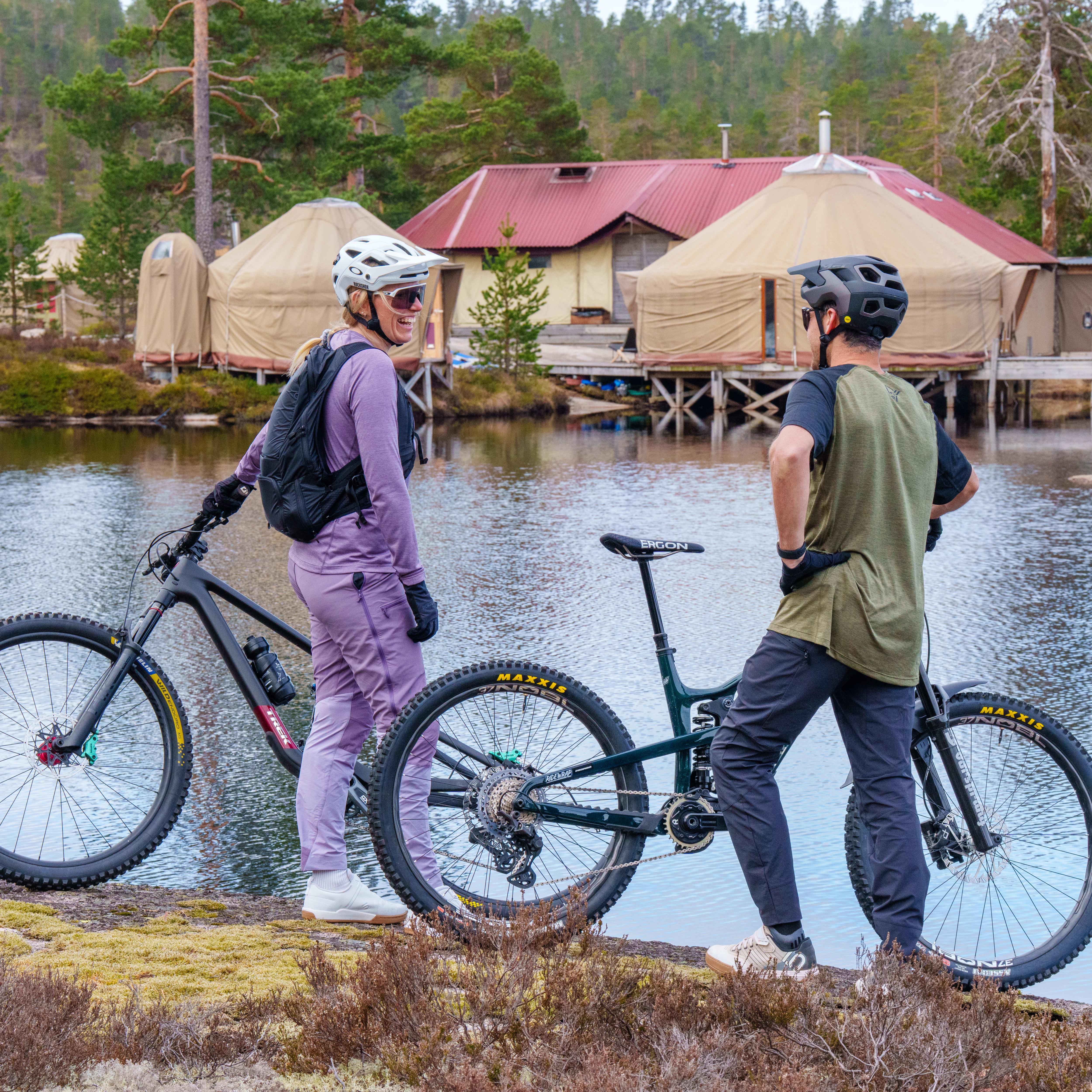 Two mountain bikers wearing Norrøna clothing stand with their bikes next to a lake with yurts behind them.