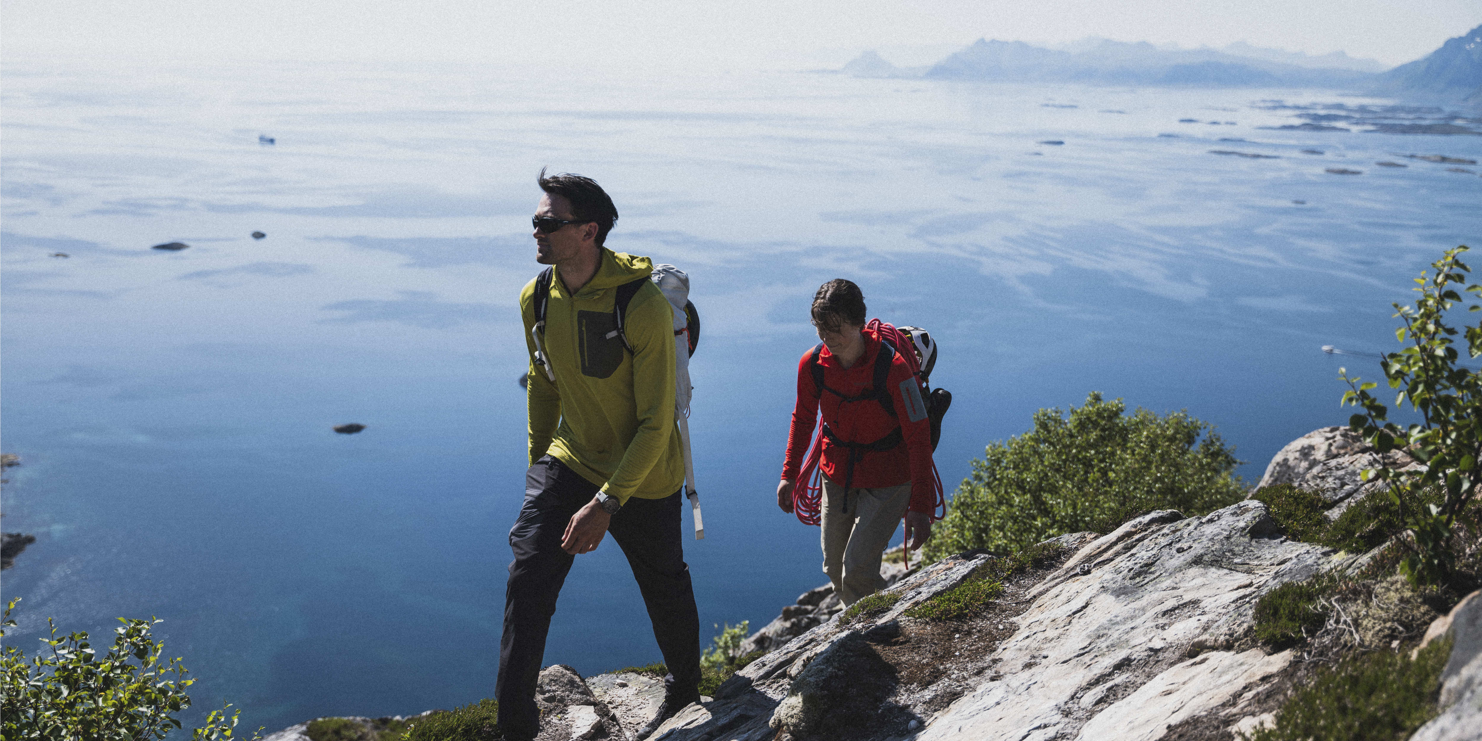 Two hikers on a rocky trail with a vast ocean view. The man wears a yellow Norrøna hooded top and sunglasses. The woman wears a red Norrøna top and carries climbing gear.