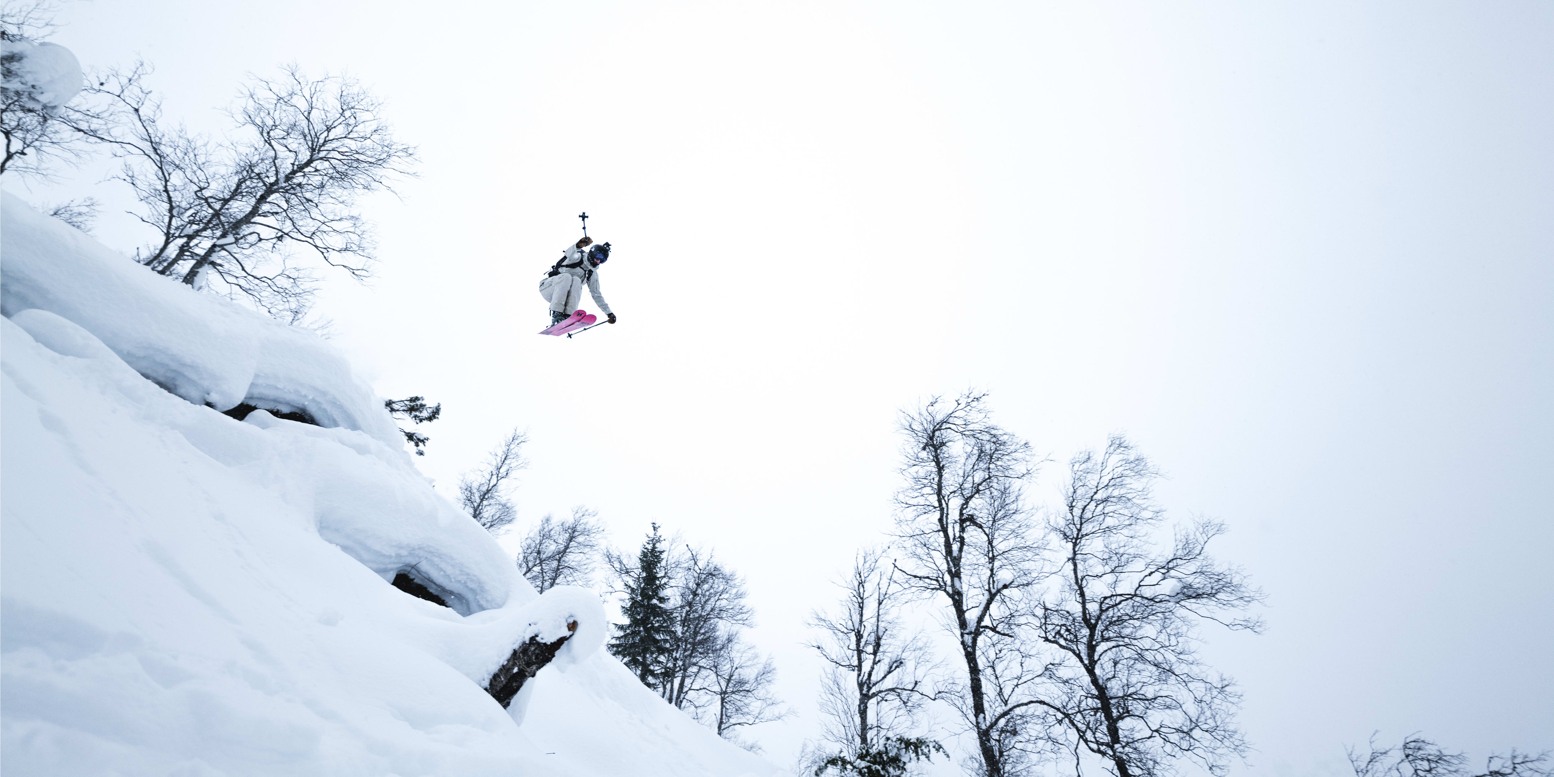 A person in a white ski suit and a black helmet is mid-air while skiing, with two black ski poles and pink skis. They are in the air above snow-covered terrain and bare trees. The sky is clear and light blue.
