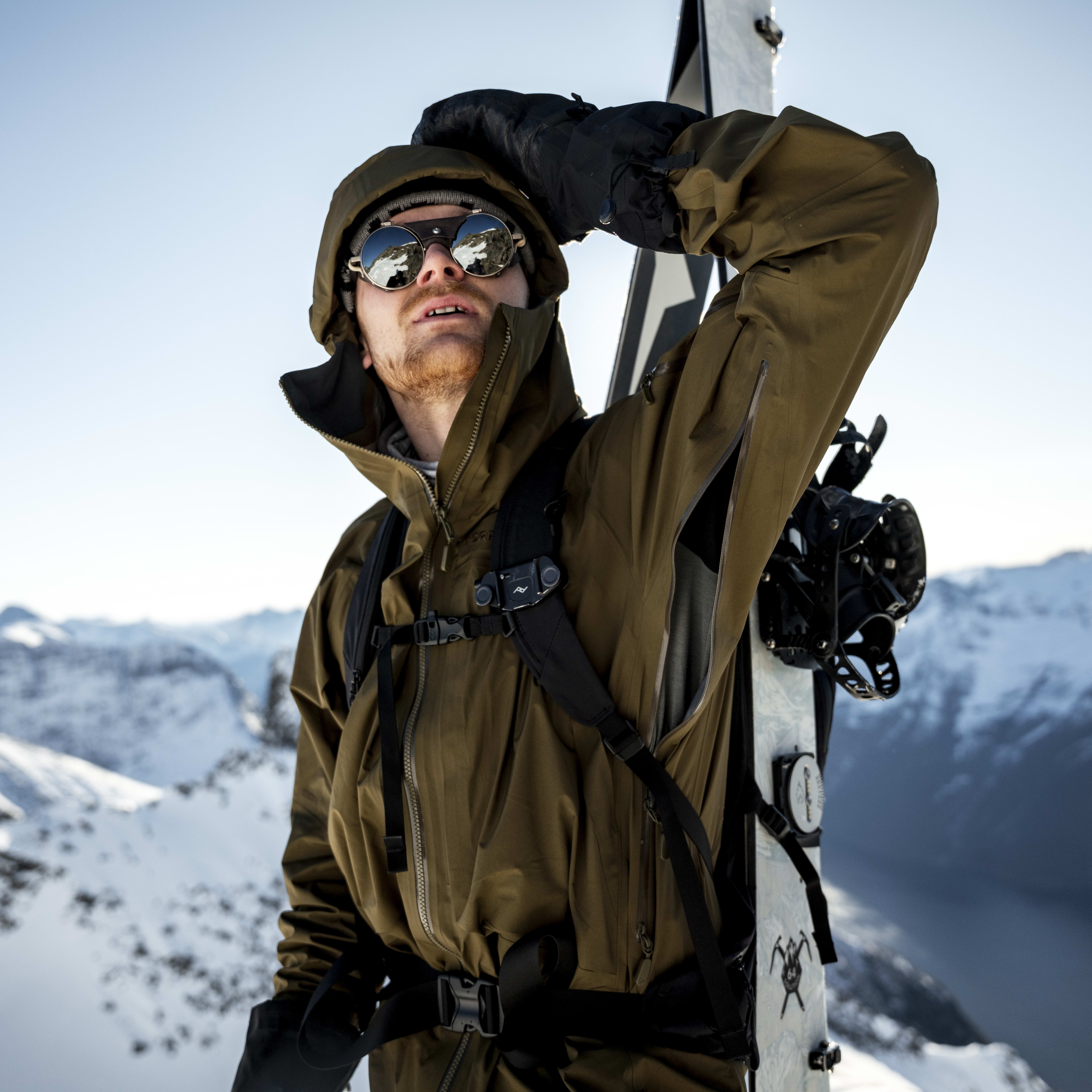A skier in a Norrøna jacket and mirrored sunglasses stands in the snow with skis on his back, gazing towards snowy mountains.