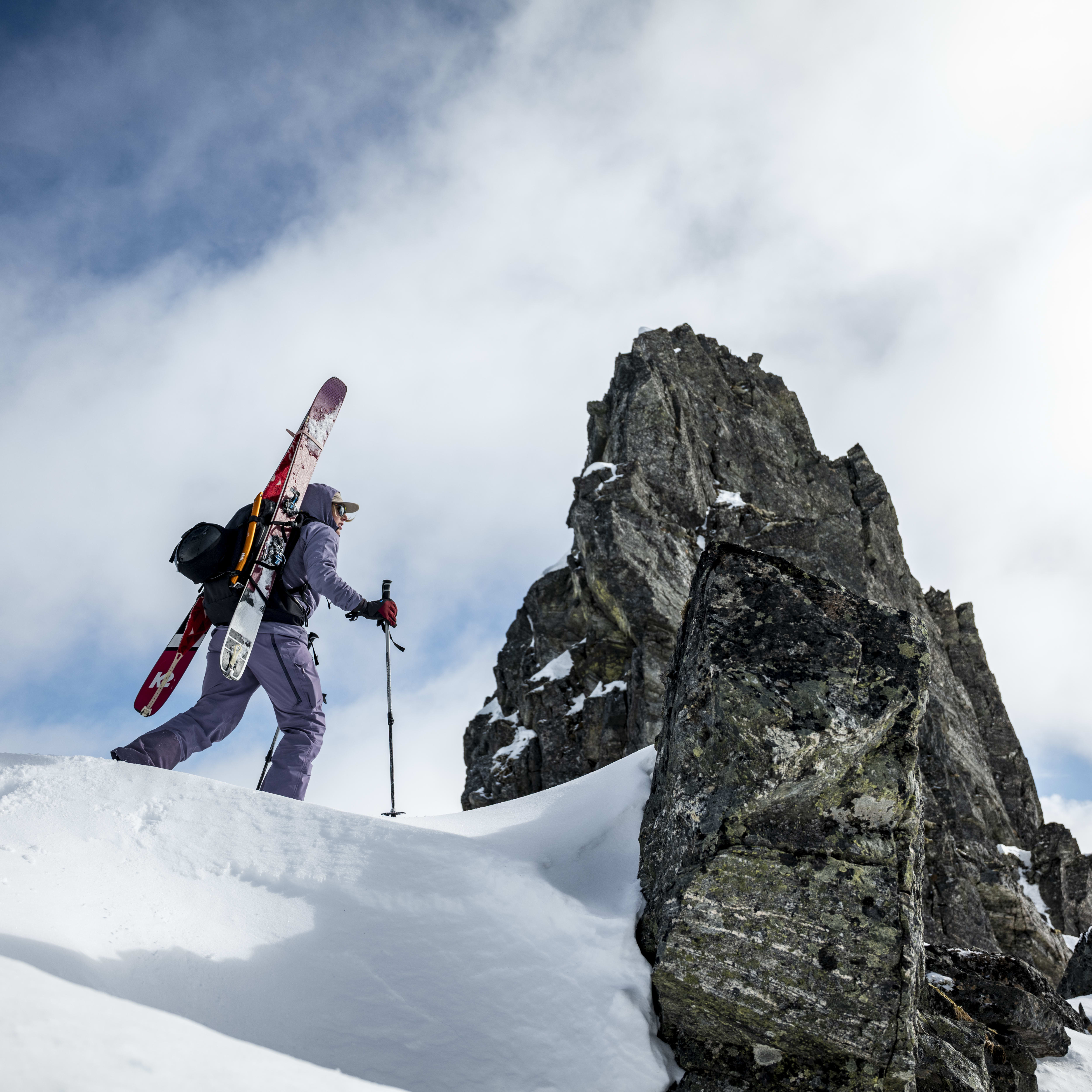 A skier with skis on their back in Norrøna clothing ascends a snowy mountain peak, using ski poles for balance. Large rock formation to the right.