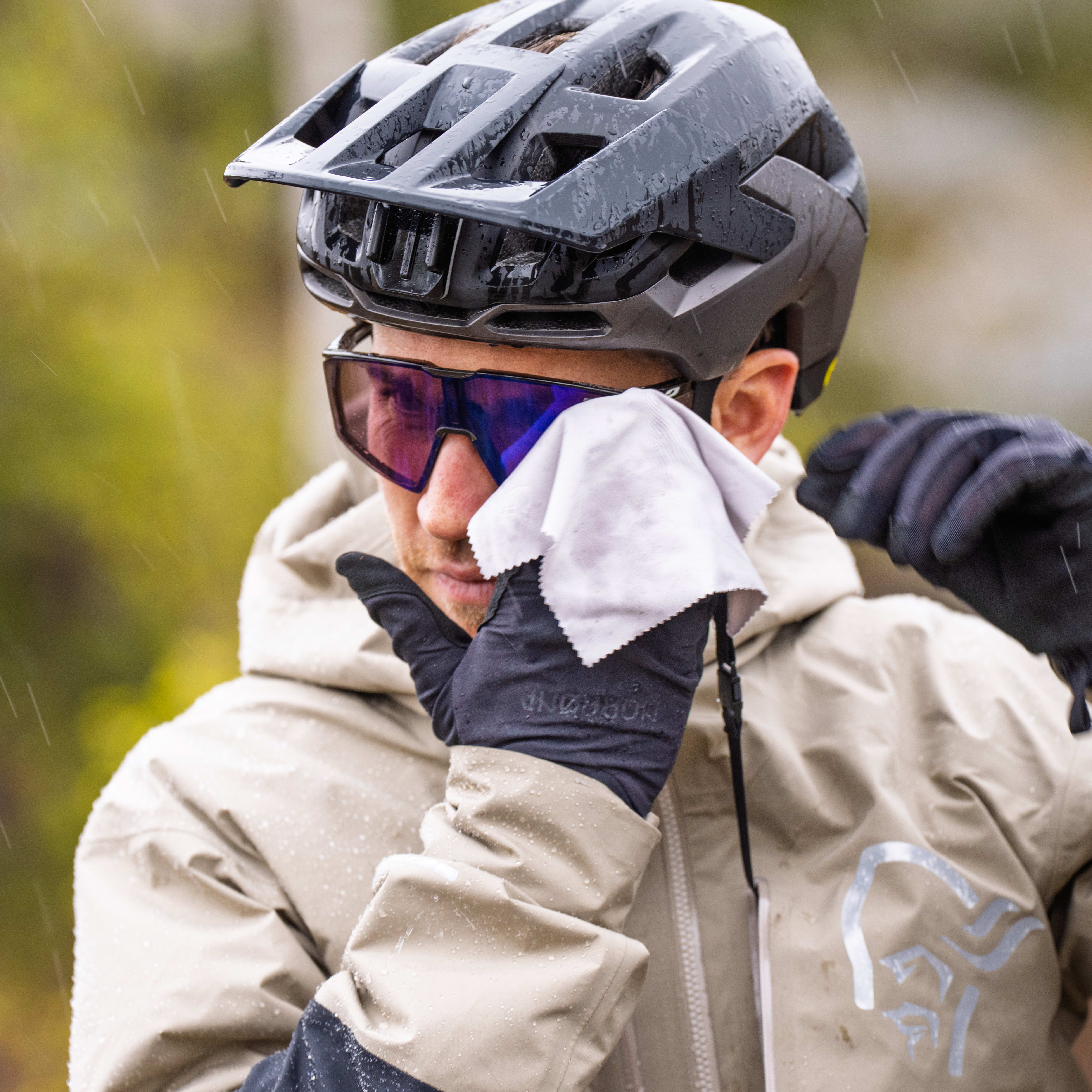 A cyclist in a Norrøna jacket wipes his sunglasses with a cloth in the rain.