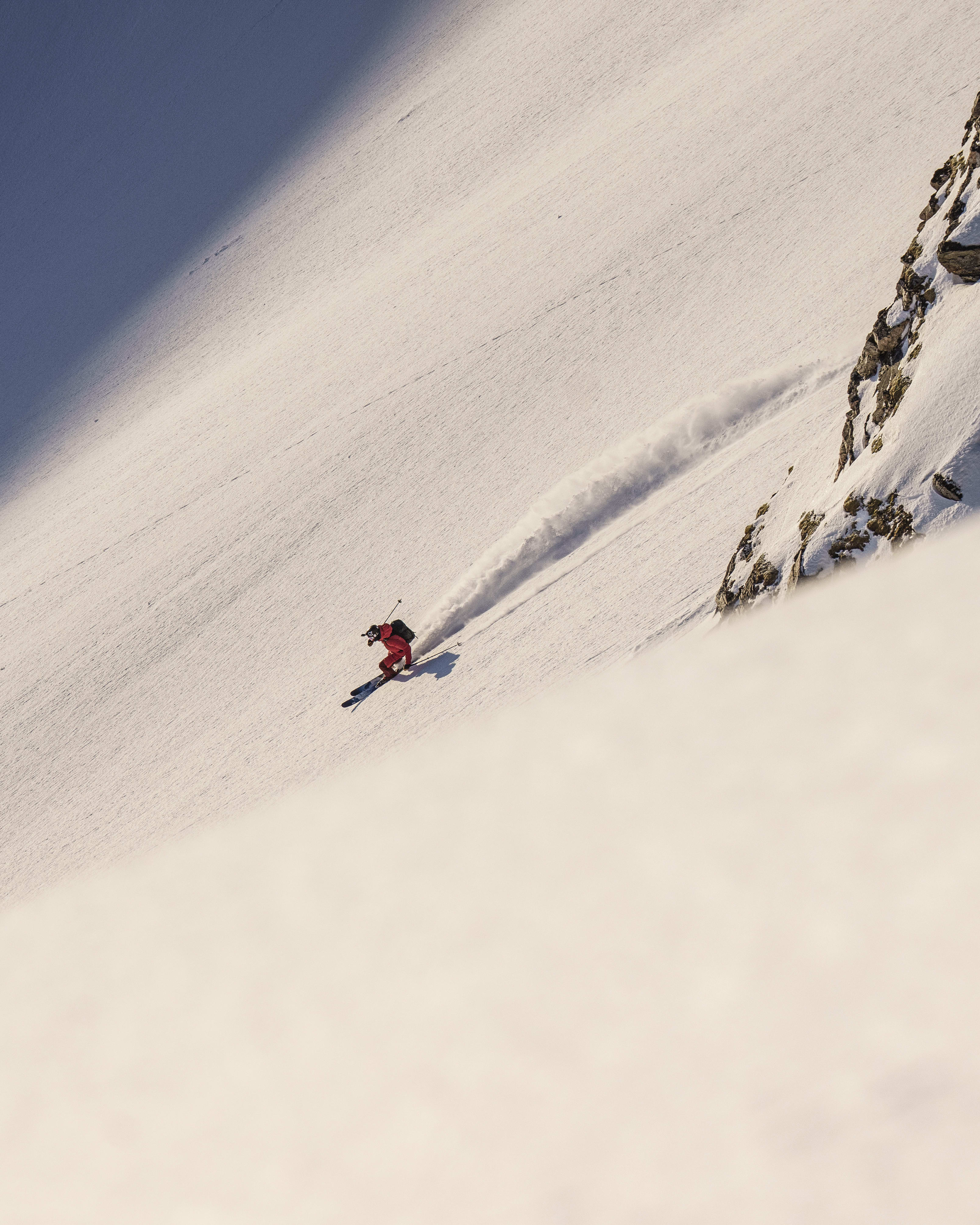 A skier in a red Norrøna suit skis down a snow-covered slope, leaving a trail of powder, with rocky terrain on one side.