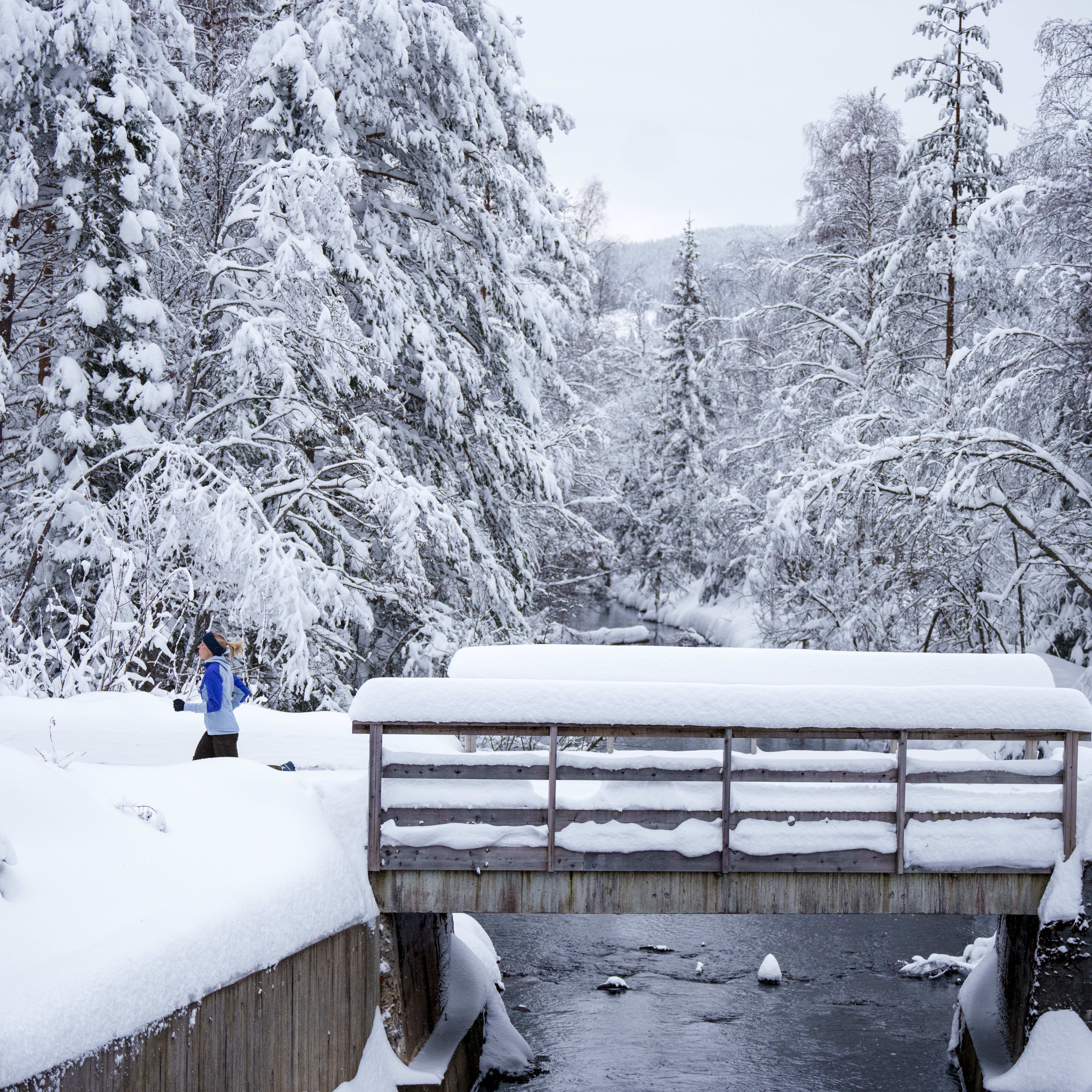 A person wearing Norrøna clothing walks across a wooden bridge covered in snow. The bridge spans a small river in a snowy forest. 
