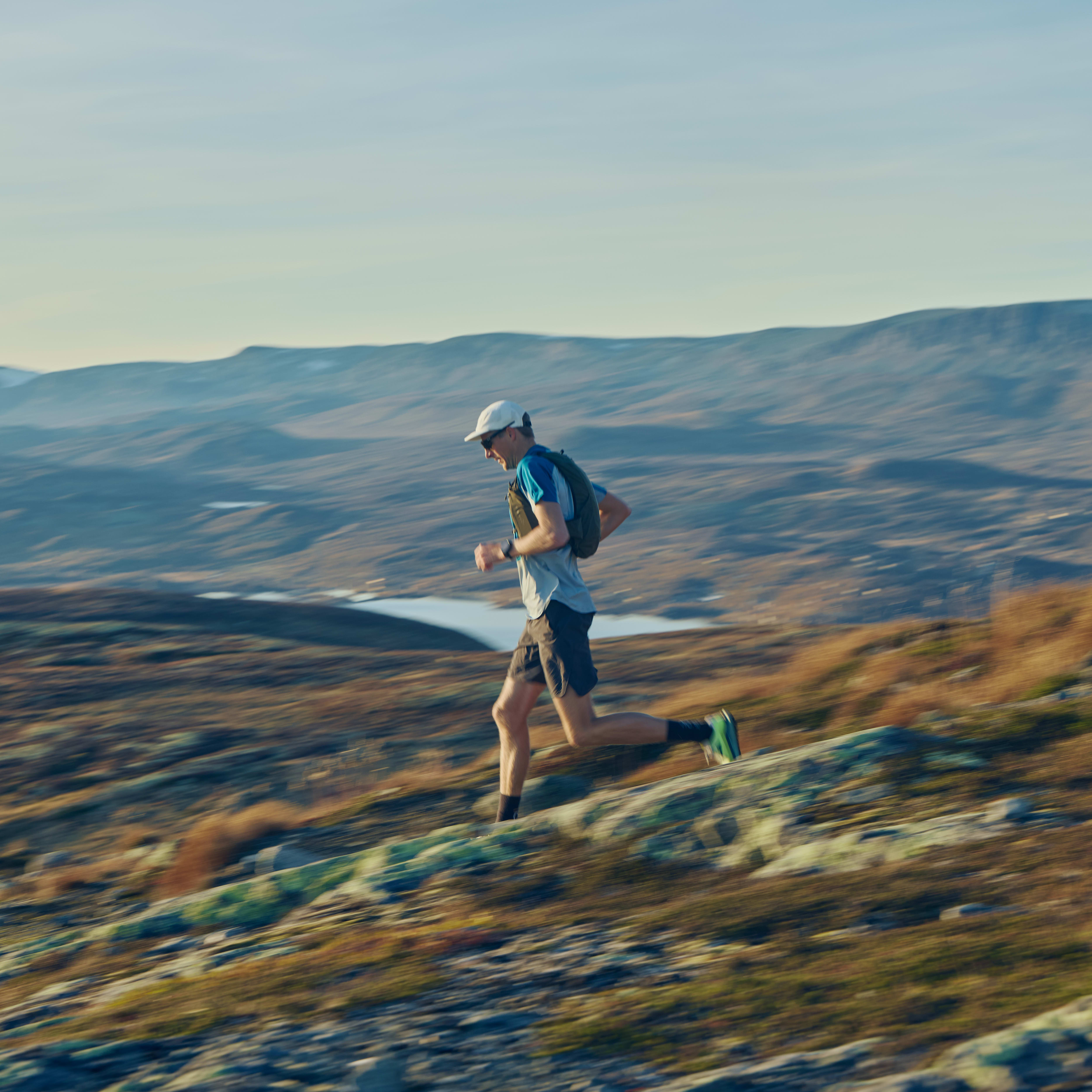 A man runs on a mountain ridge, wearing Norrøna shorts and a hydration pack, with mountains and a lake in the background.