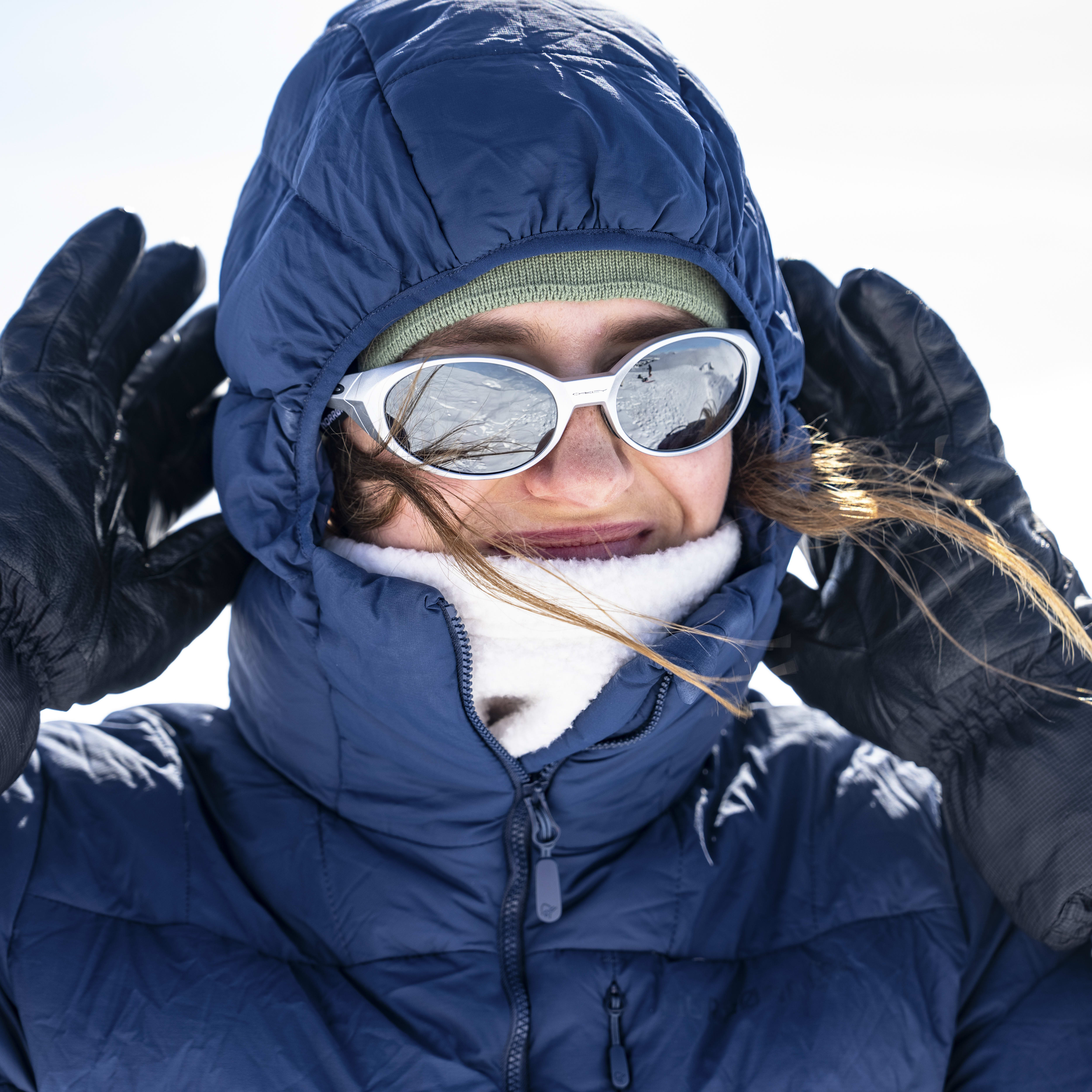 A woman wearing a blue Norrøna jacket and black gloves with a white Gore-Tex logo. The jacket is zipped up and the woman is wearing sunglasses.