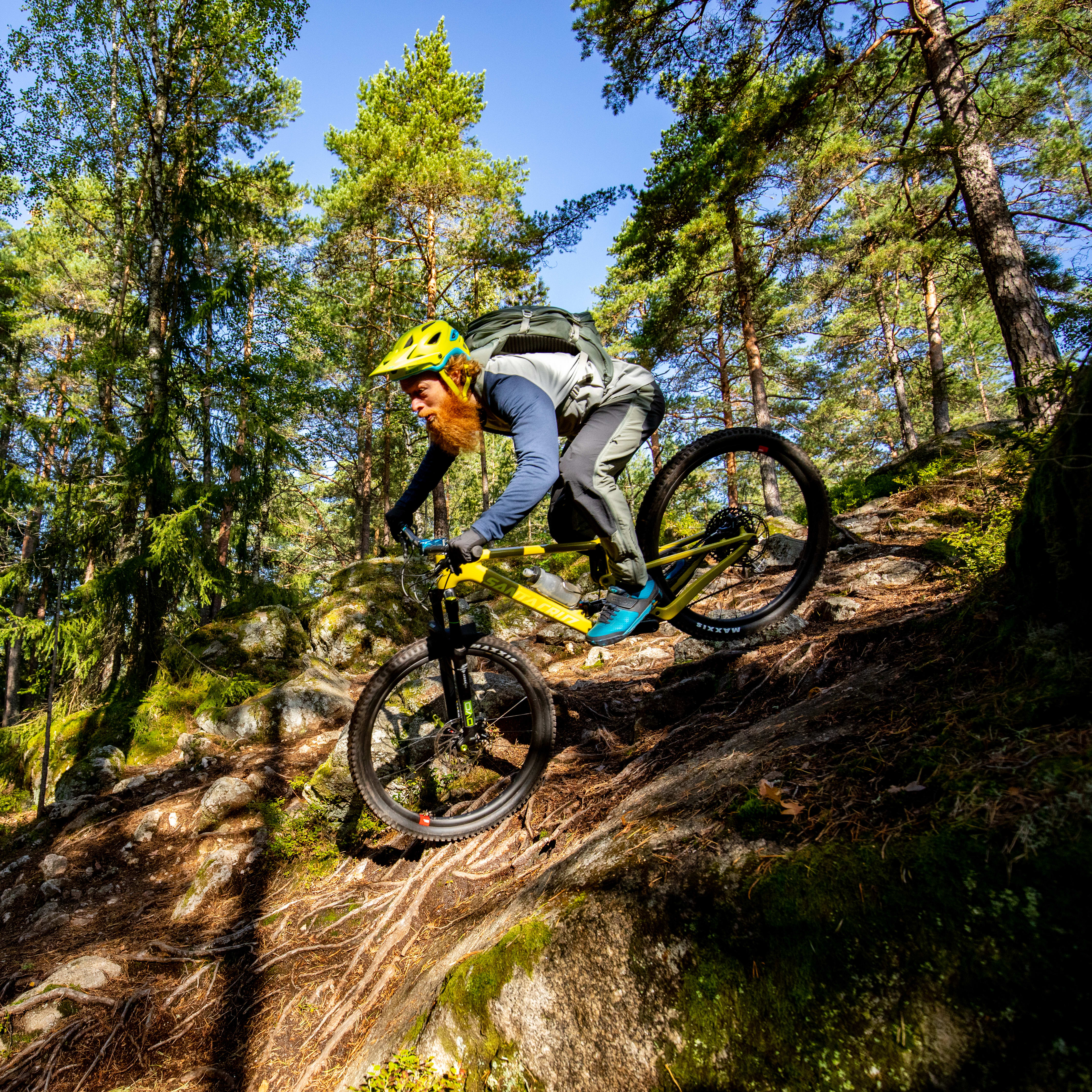 A man wearing a Norrøna jacket rides a mountain bike on a rocky trail through a forest. 

