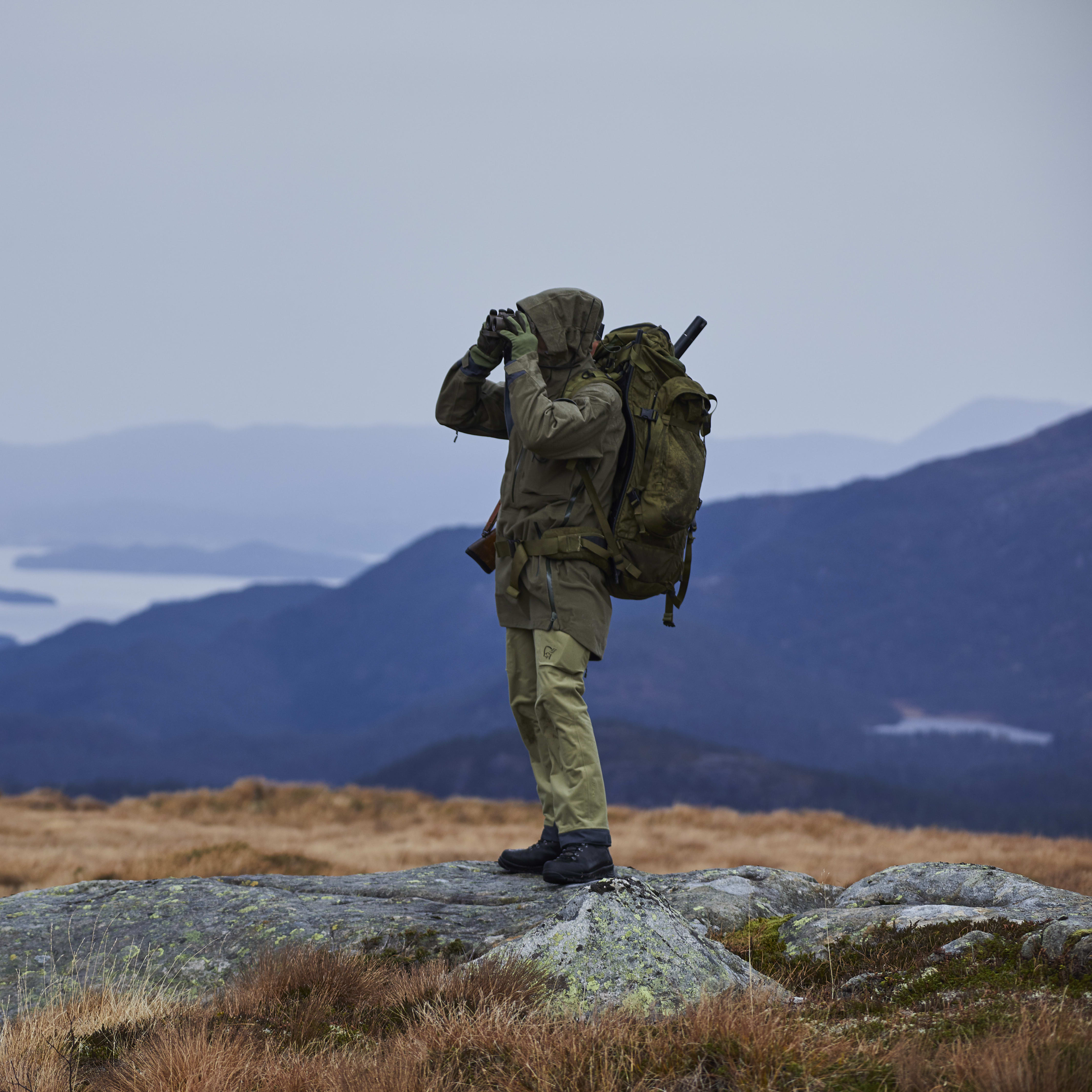 A person wearing a Norrøna jacket and pants stands on a rock in a mountain landscape.  
