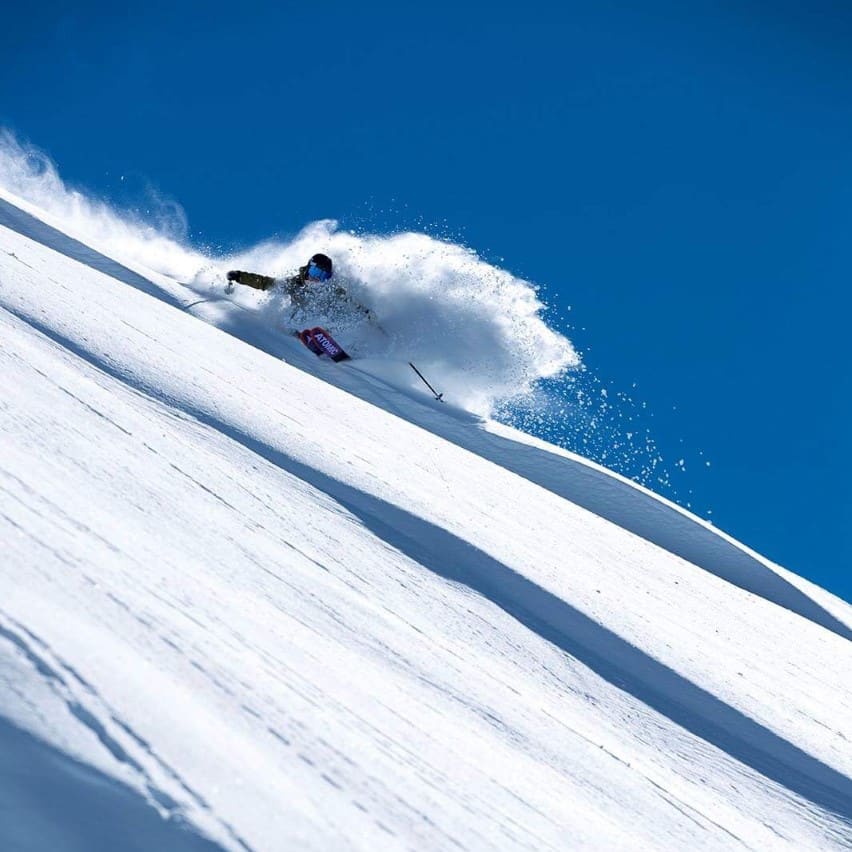 A skier in Norrøna gear carves down a steep, snow-covered slope against a bright blue sky, kicking up a cloud of powder.
