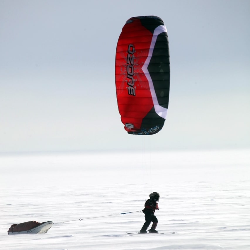A person in Norrøna apparel snow kites with a red and black Ozone kite and pulls a sled across a snowy field on a bright day.
