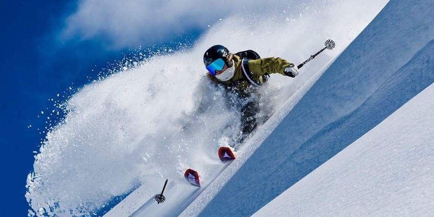 A skier in Norrøna clothing carves through deep powder on a sunny, snowy slope with blue sky visible.