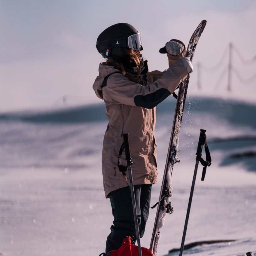 A skier in a helmet and Norrøna jacket preps skis on a snowy landscape. Ski poles and a red backpack rest nearby.
