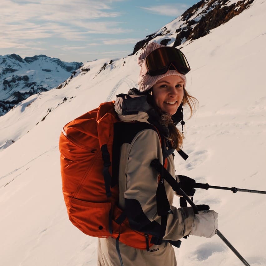 A smiling skier in the mountains, wearing a pink hat, goggles, and a tan Norrøna jacket with an orange backpack, holding ski poles.

