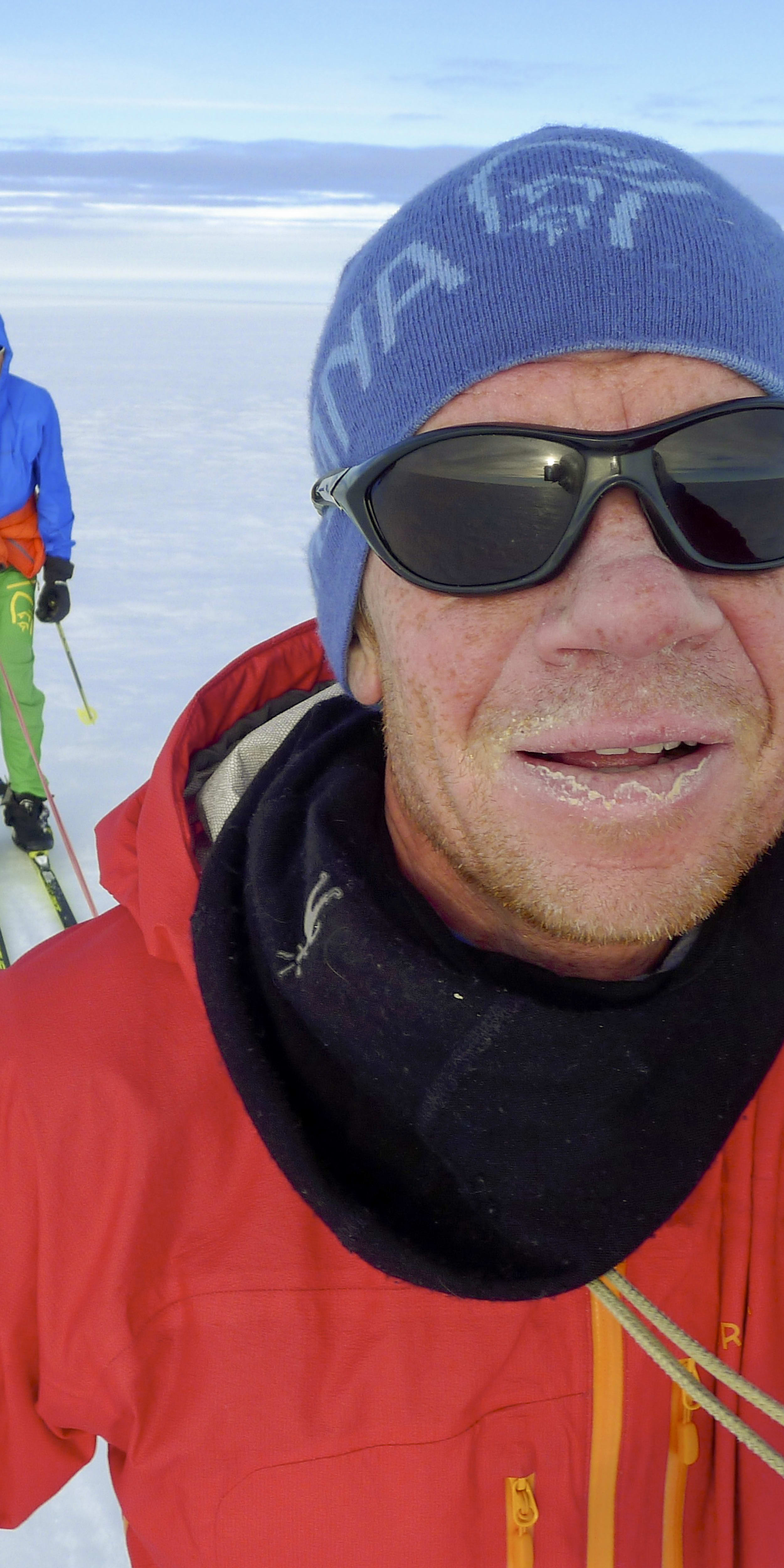 A person in a red jacket and black sunglasses takes a selfie while cross-country skiing. Two other people cross-country ski behind them, pulling sleds. The sky is bright blue with white clouds and the ground is covered in snow.