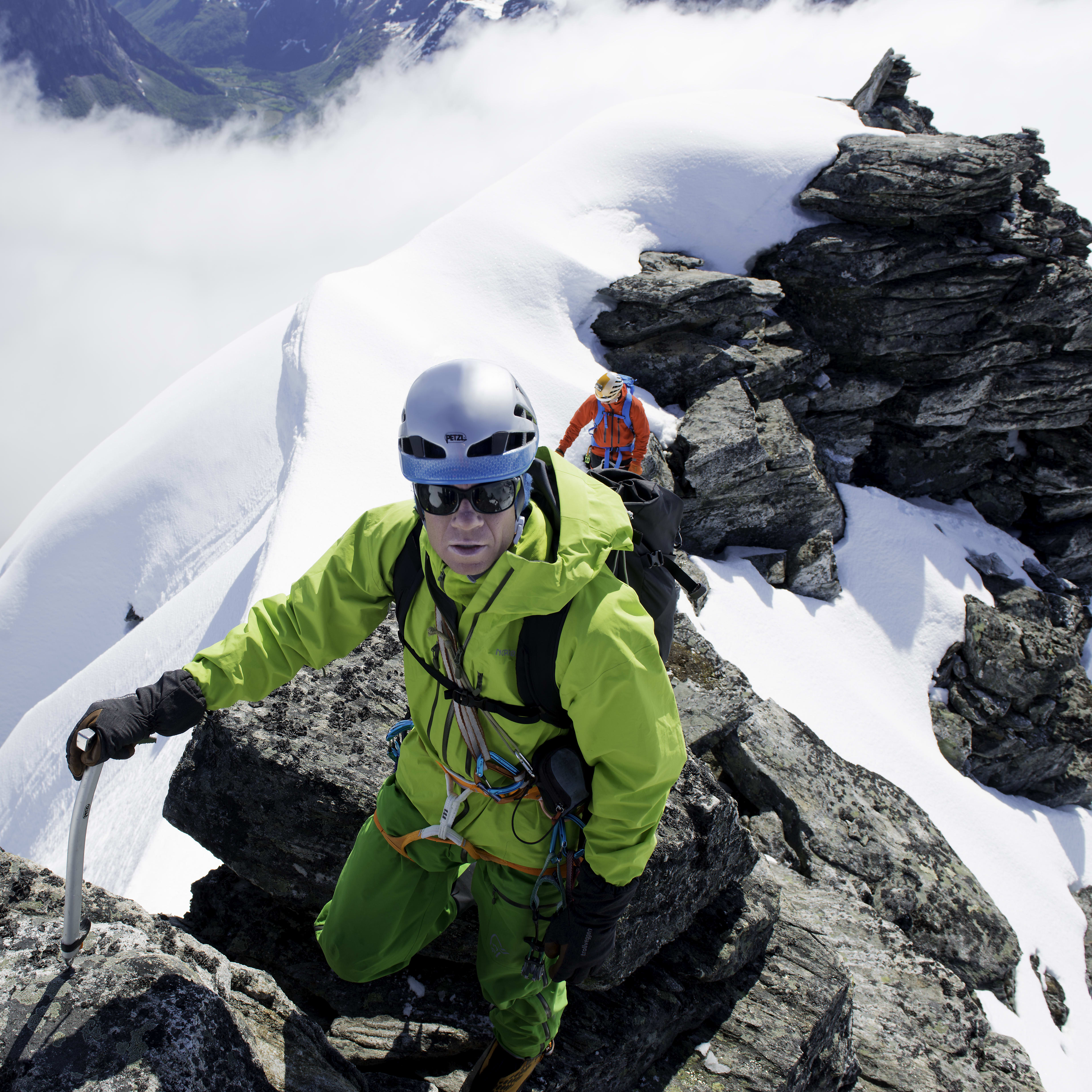 A climber wearing a Norrøna jacket and helmet is on a snowy mountain peak. He's holding onto an ice axe. The view behind him is a snowy valley.