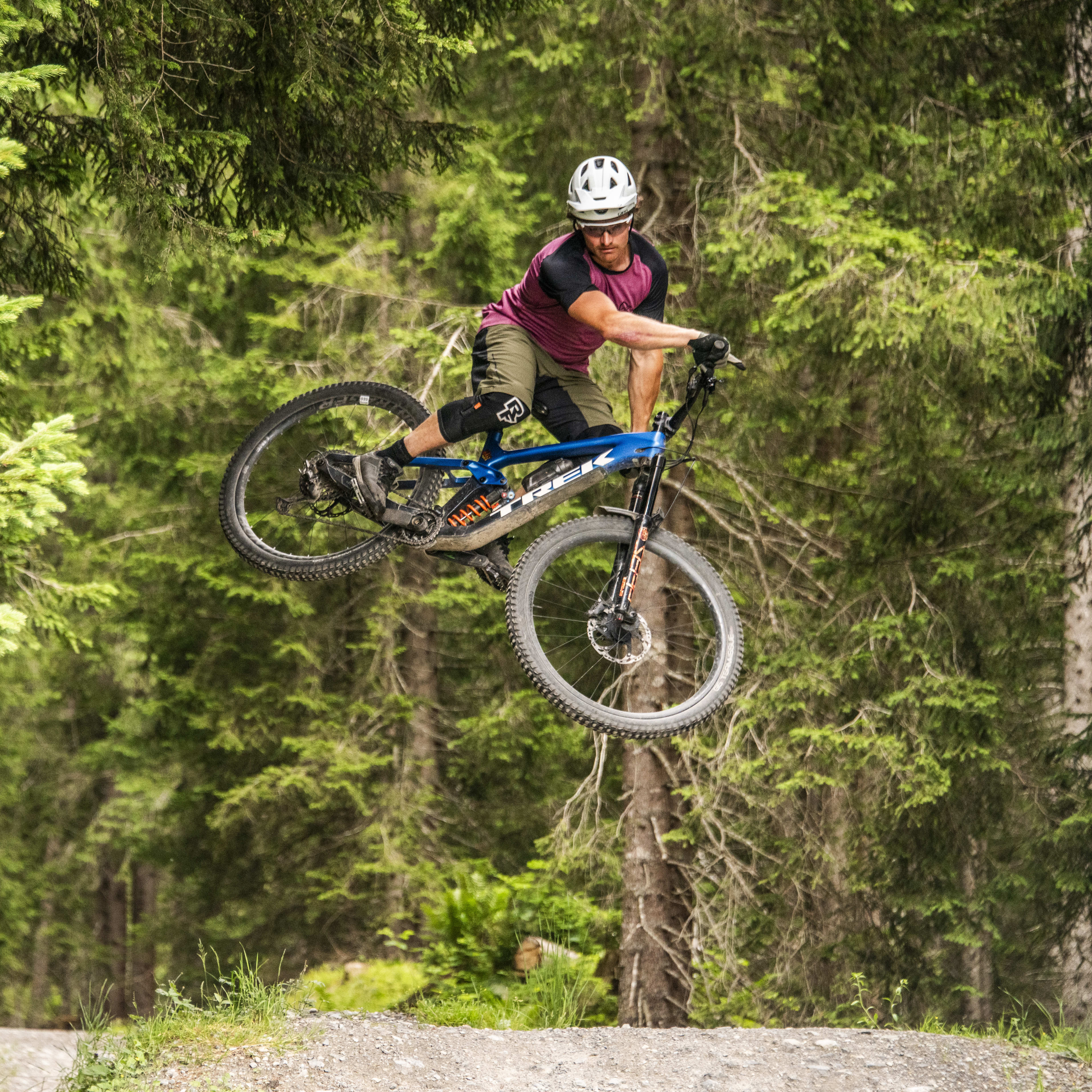 A man in a Norrøna shirt jumps a mountain bike over a dirt trail in a forest.