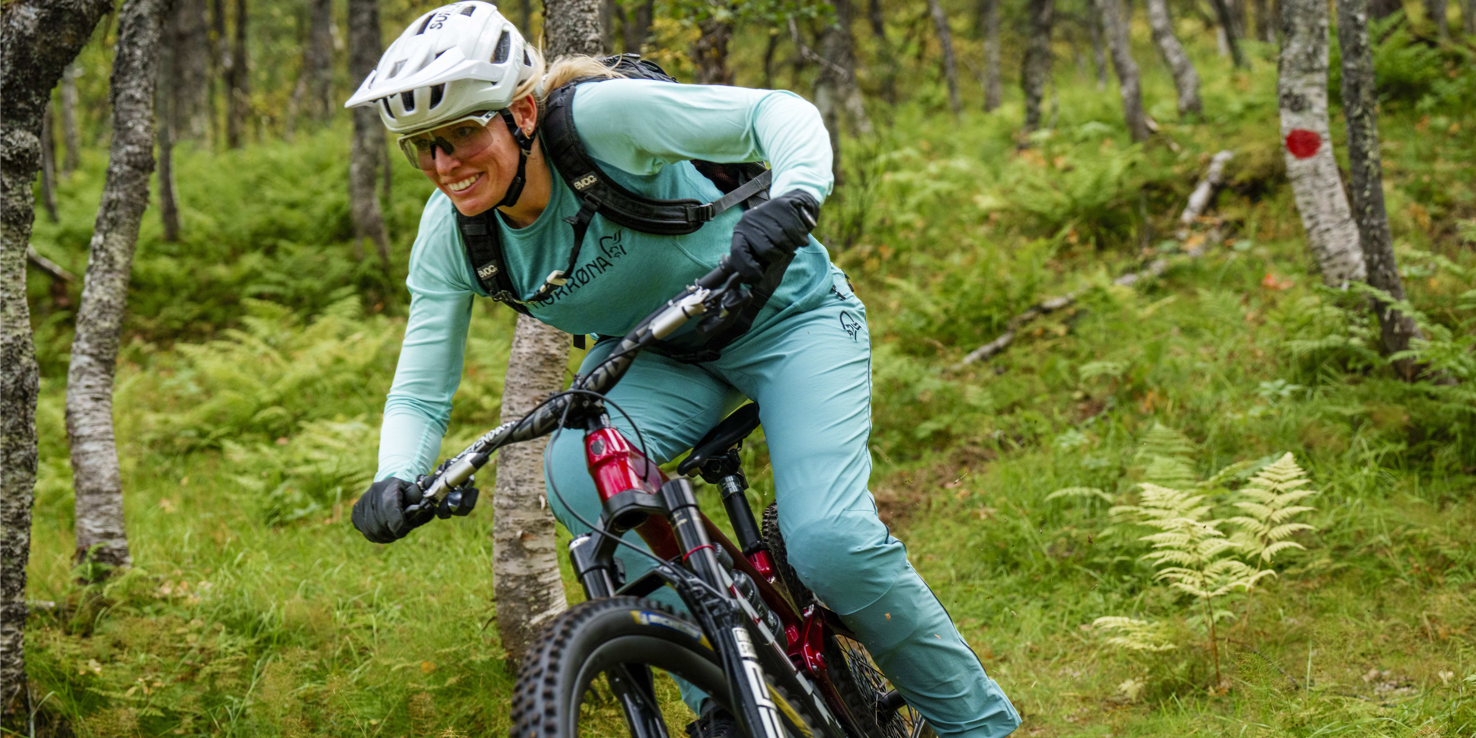 A smiling woman in Norrøna teal clothing mountain bikes through a forest. She wears a helmet and sunglasses.
