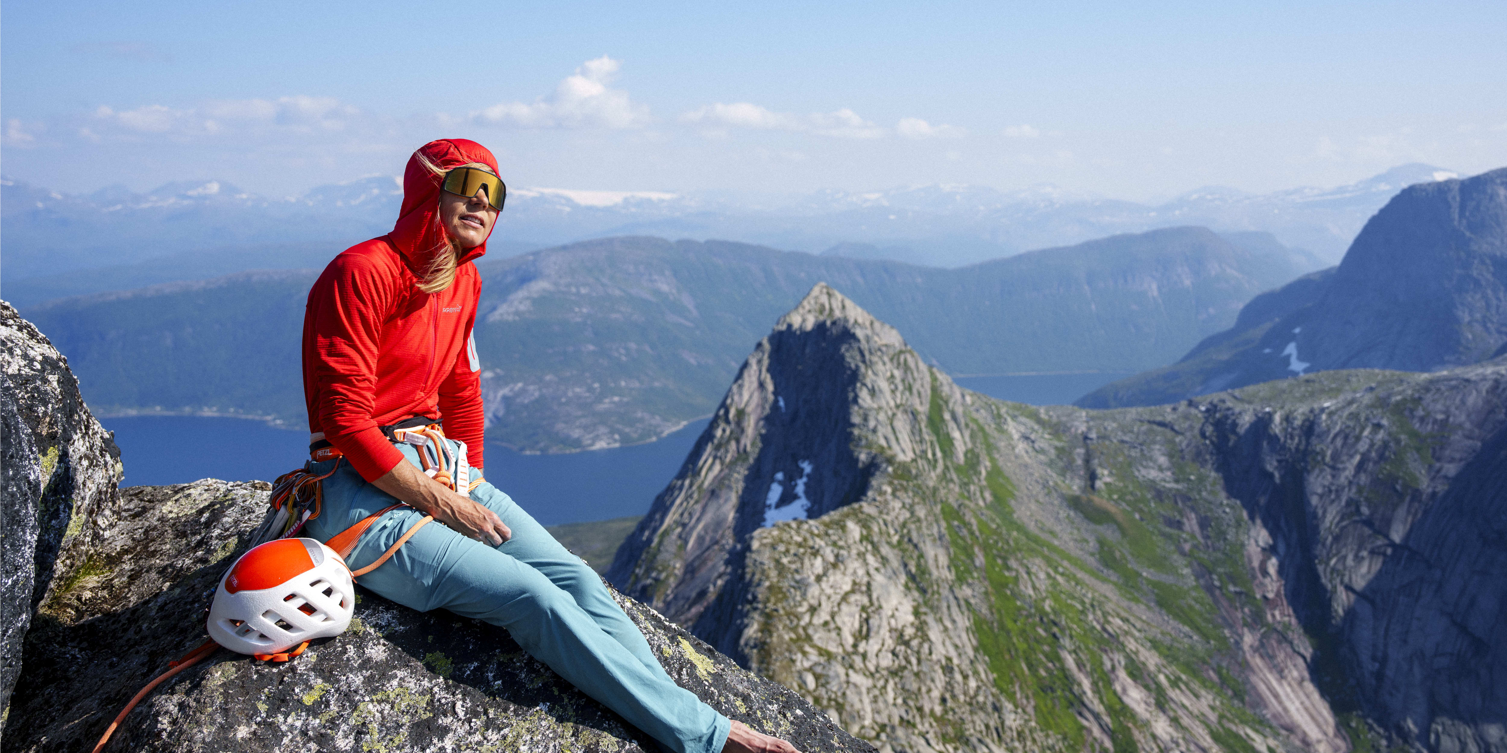 A climber wearing a red Norrøna hoody and green pants sits on a rock ledge with a helmet, overlooking a mountain range under a clear blue sky.