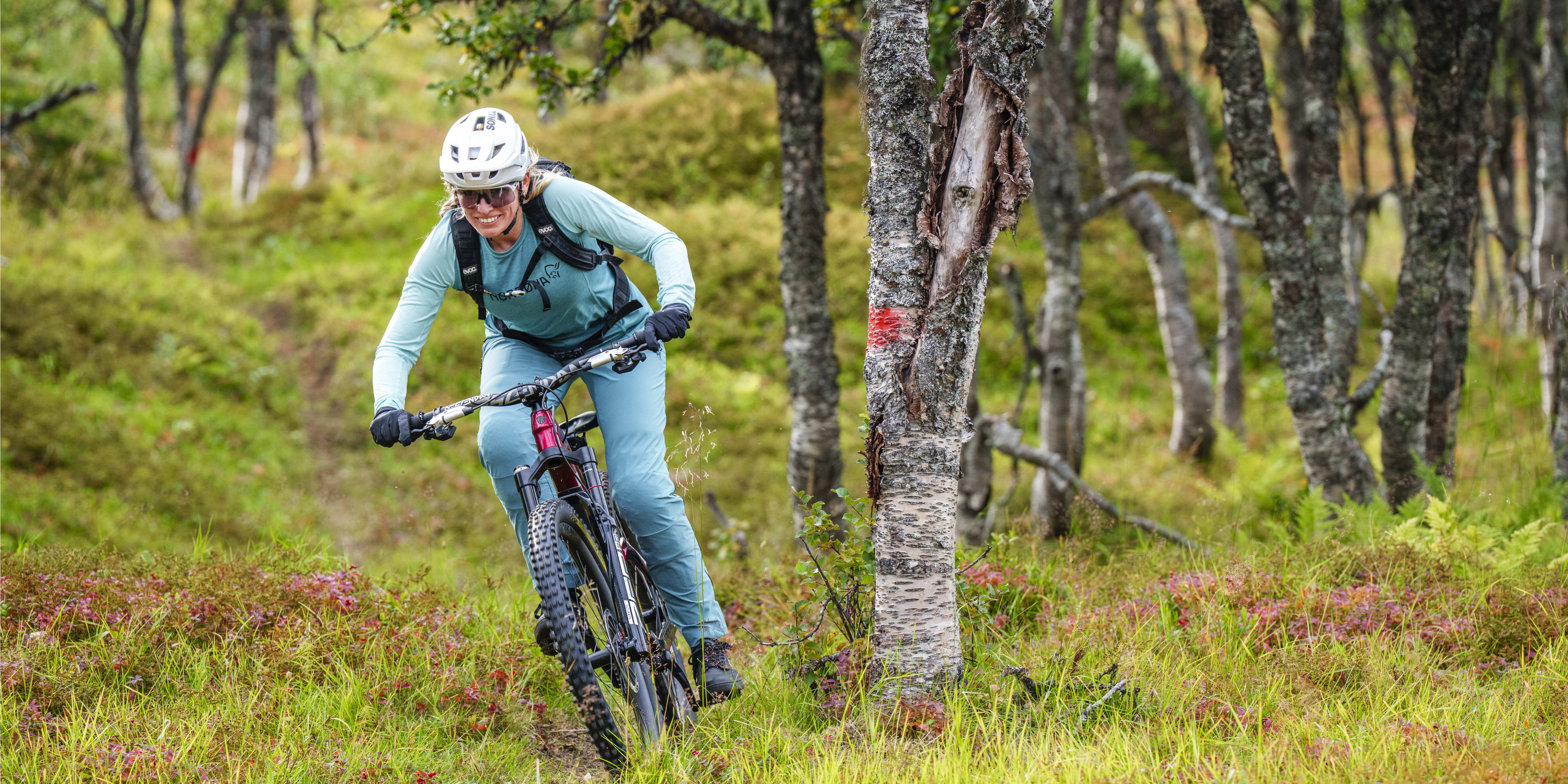 Woman mountain biking through a grassy, wooded area. She wears a helmet, sunglasses, and Norrøna apparel.