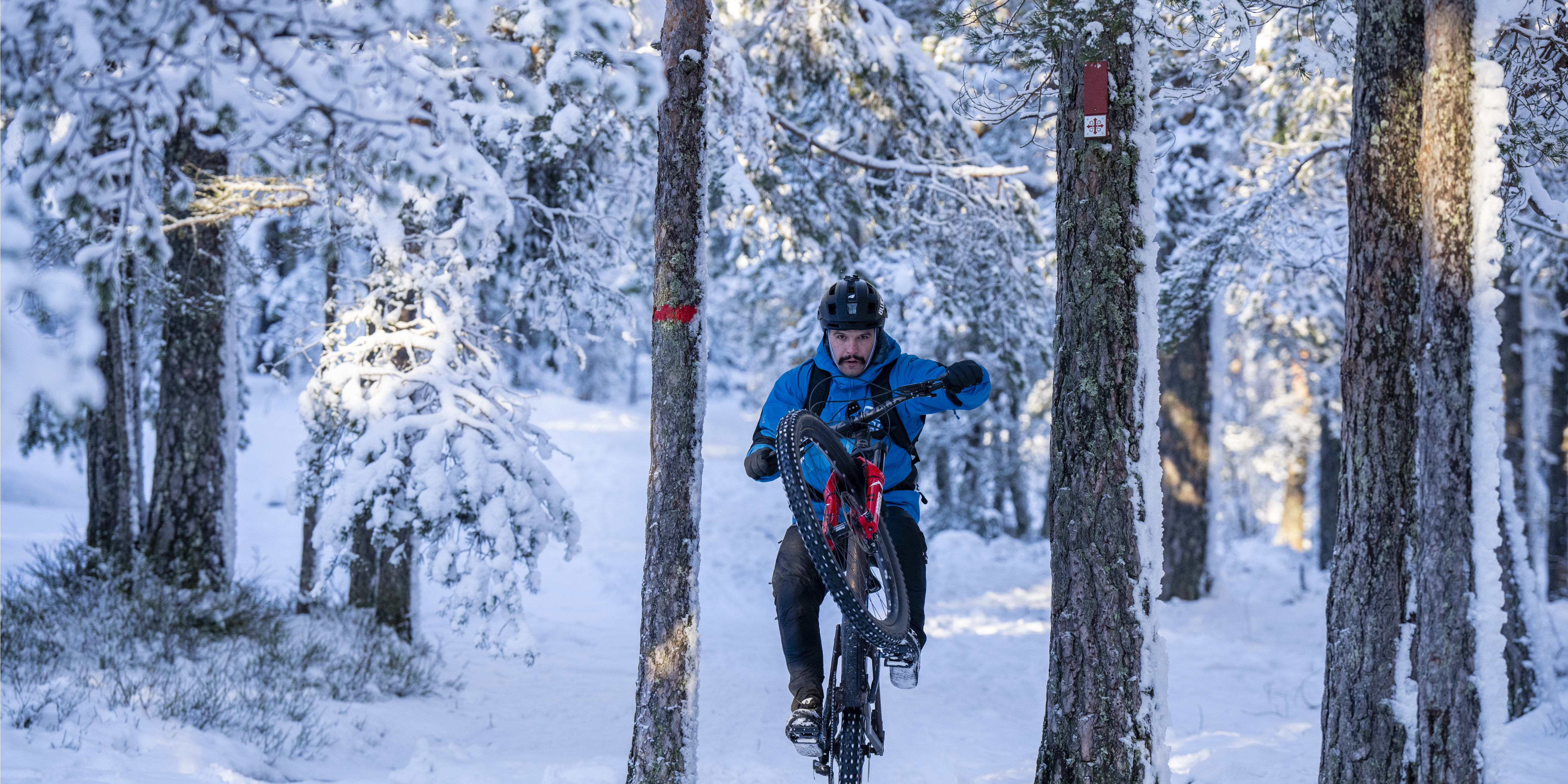 A man in a Norrøna jacket and helmet riding a mountain bike on a snowy trail in a forest. 
