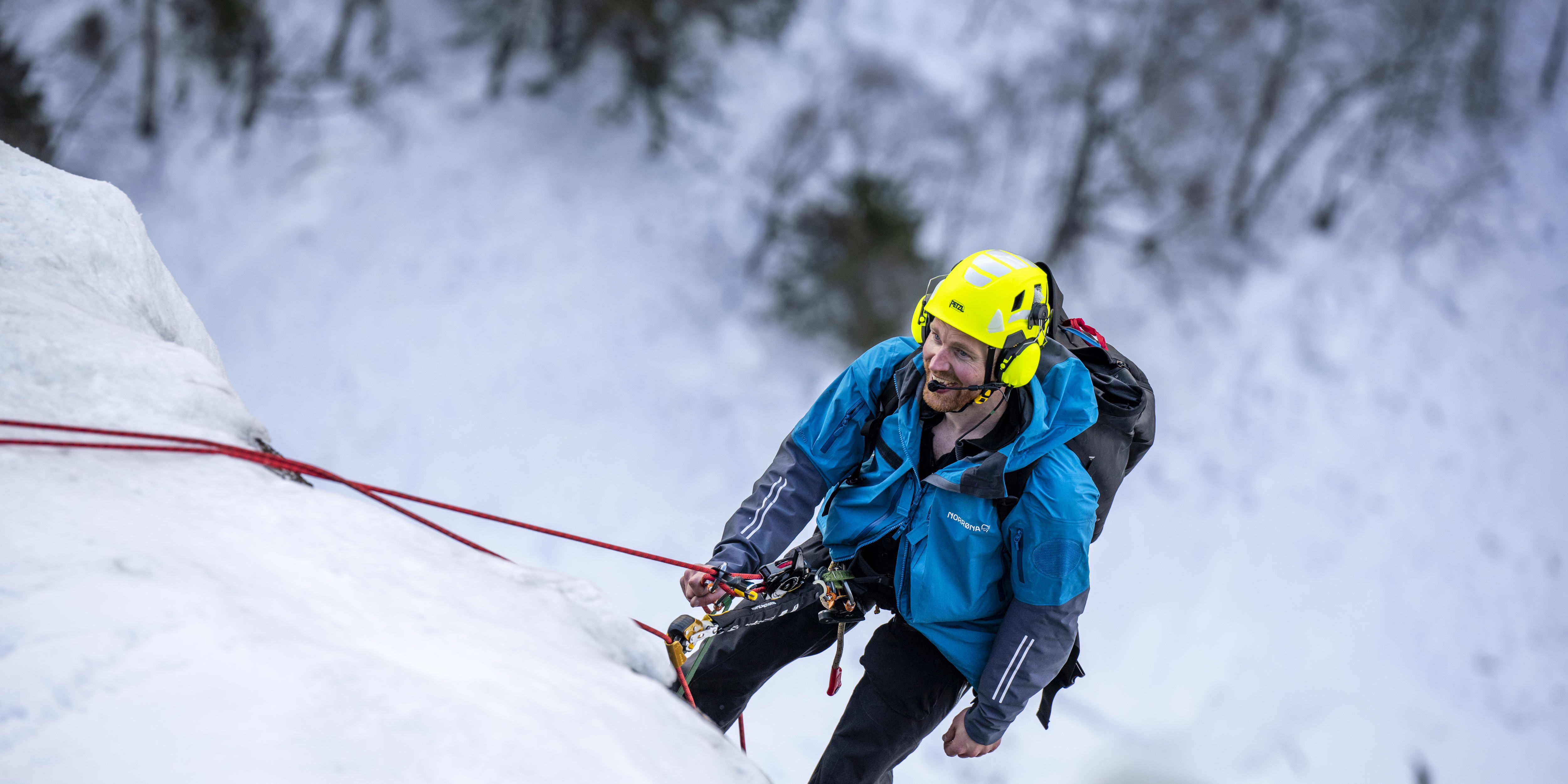 A man wearing a Norrøna jacket and a helmet climbs an icy wall.  A red rope secures him to the mountain. 
