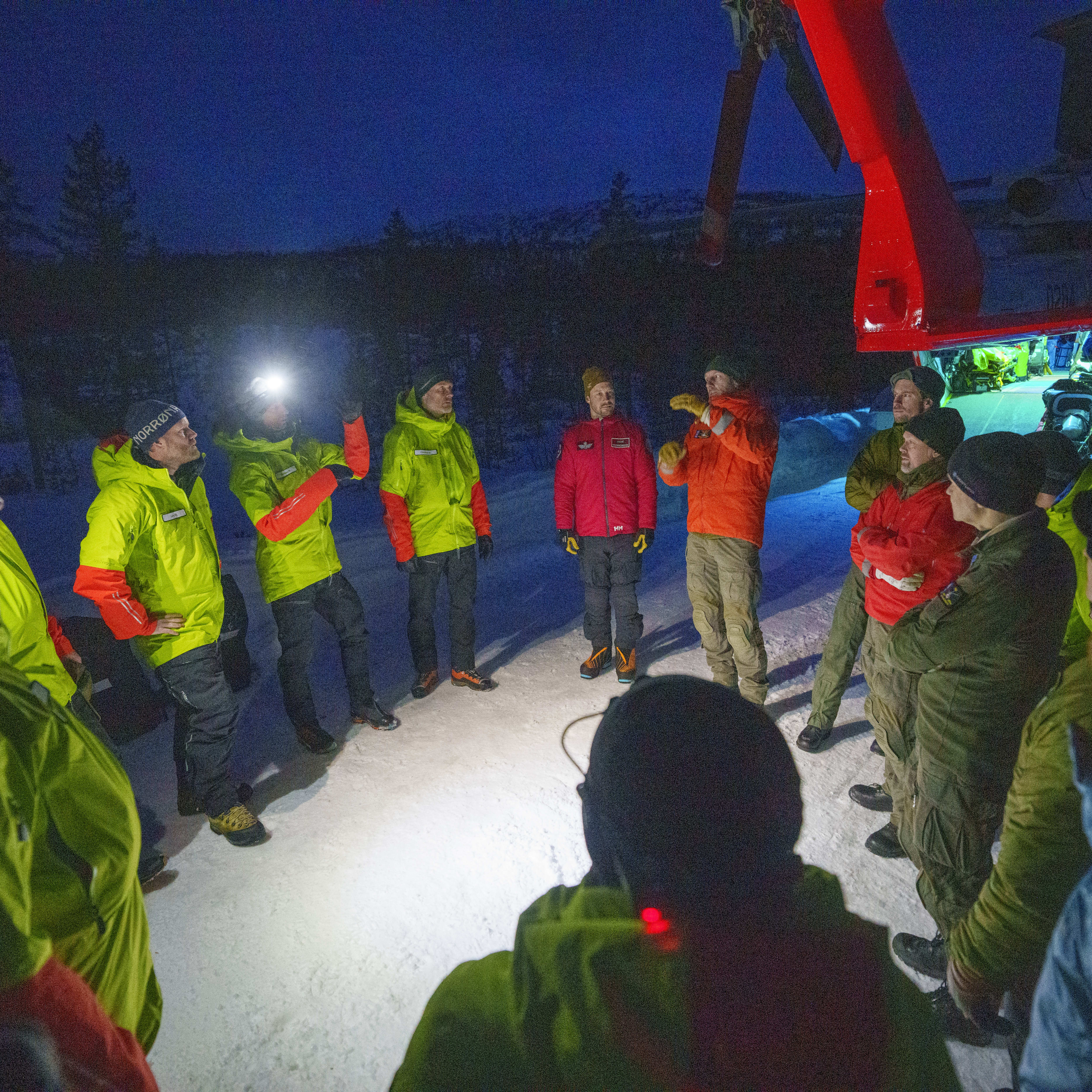 A group of people wearing Norrøna jackets stand in a snowy field with a helicopter in the background. 
