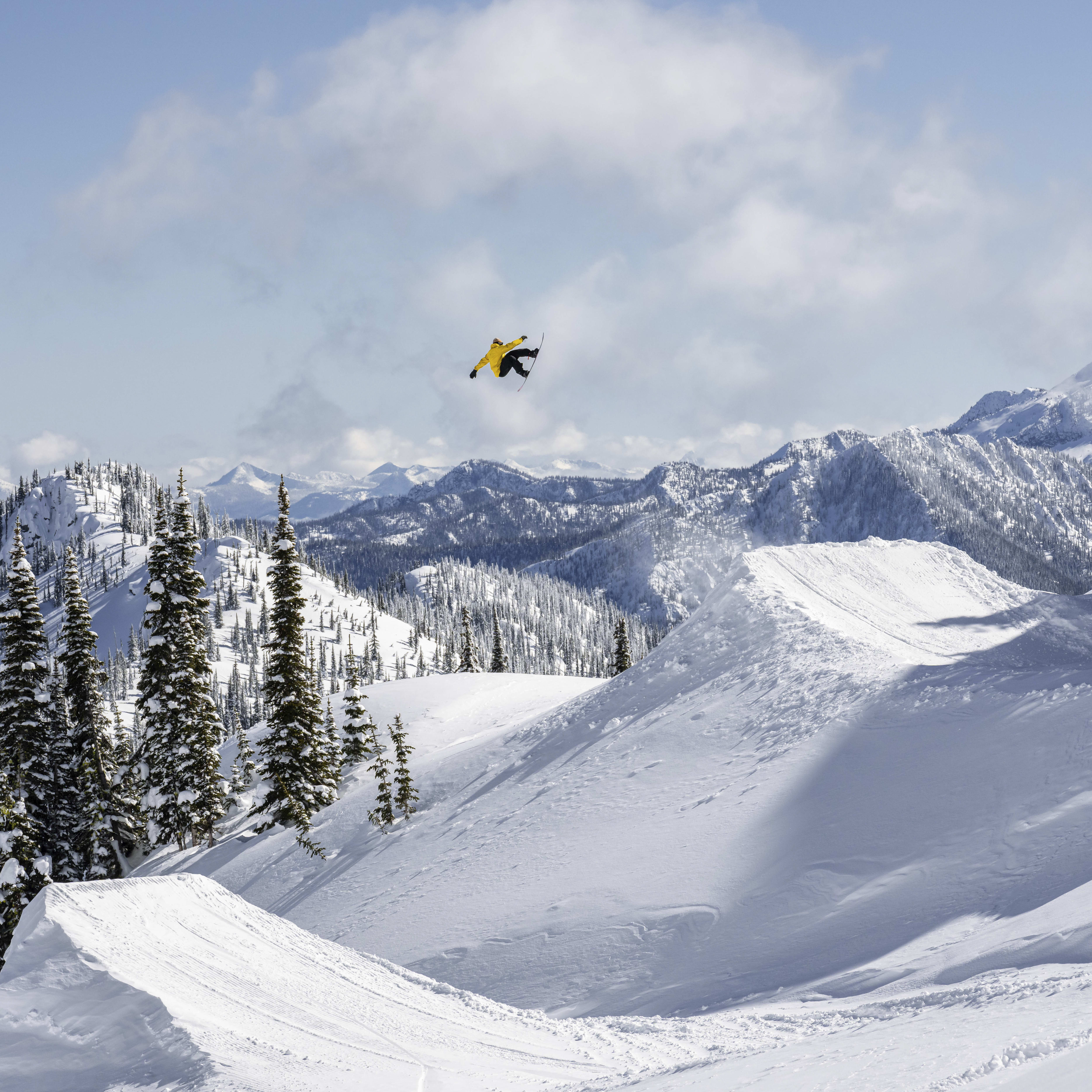 A snowboarder in a yellow jacket is mid-air over a snow-covered mountainside. The snowboarder is doing a trick. The background shows a snow-covered mountain range under a bright blue sky.