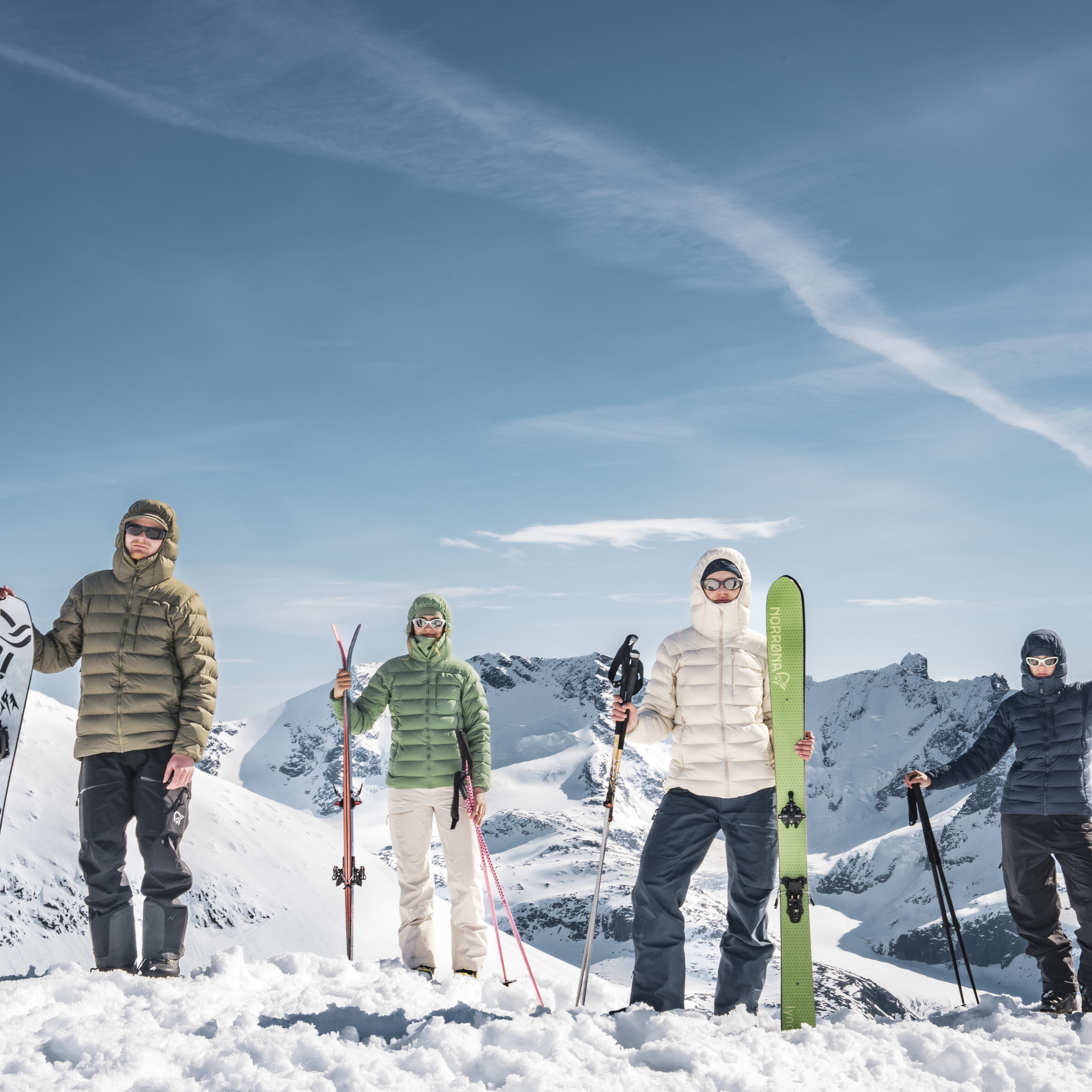 Four people in Norrøna clothing stand on a snowy mountaintop, holding skis and a snowboard.  The sky is blue with white clouds. 

