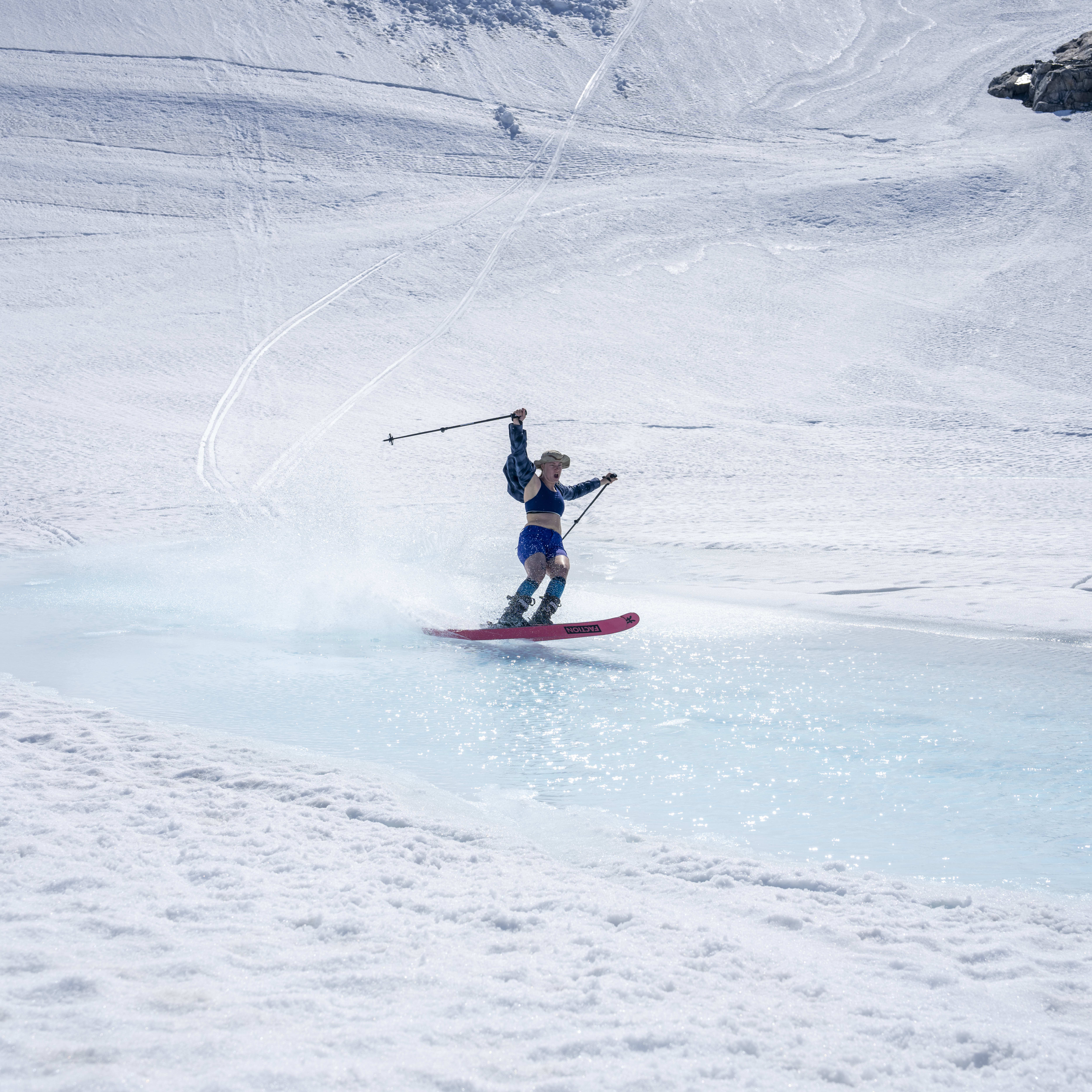 A skier wearing Norrøna clothing is performing a trick on a snowy slope. The skier is in mid-air with their skis pointed toward the water.
