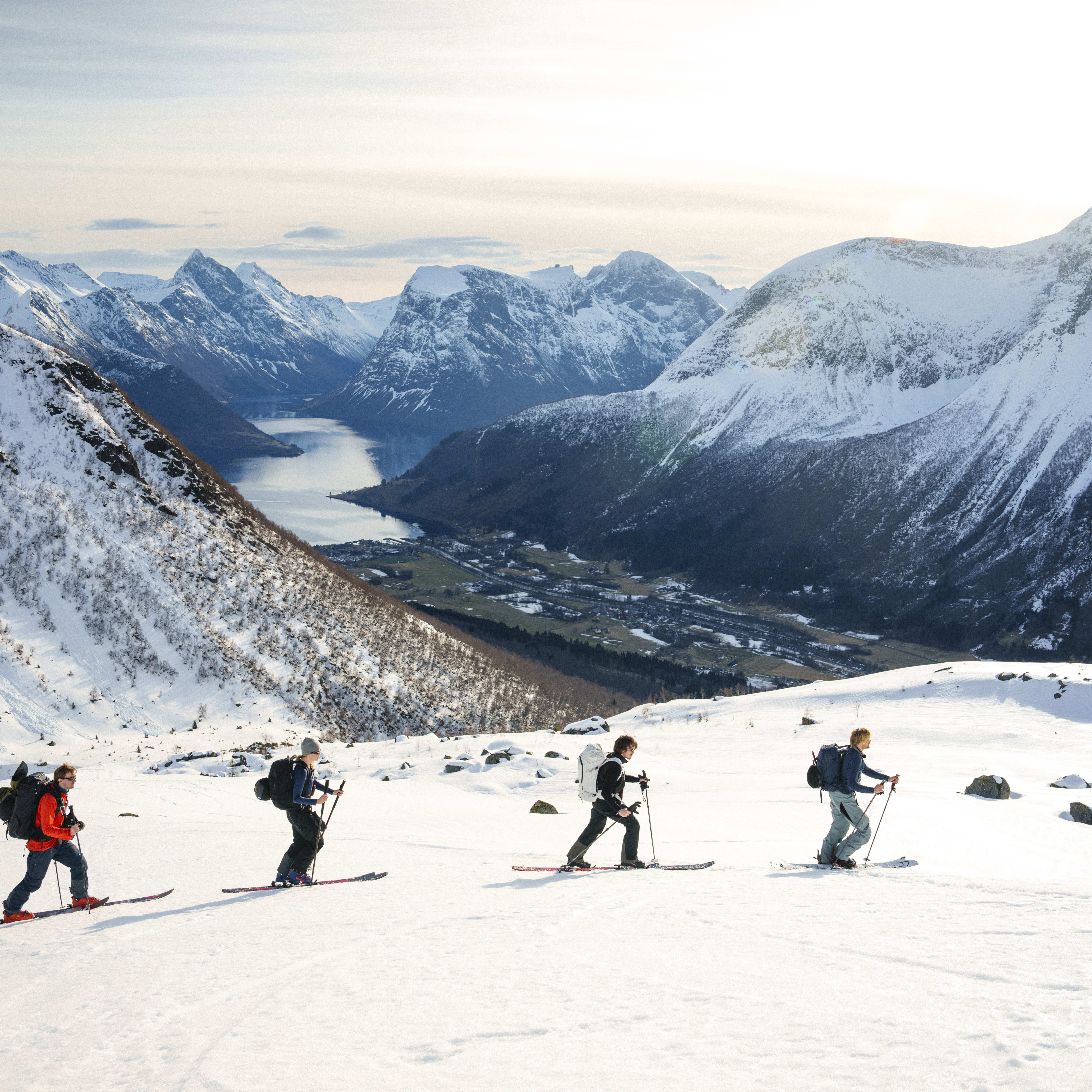 Four people ski down a snowy mountain with a fjord in the background. They are wearing Norrøna gear.  
