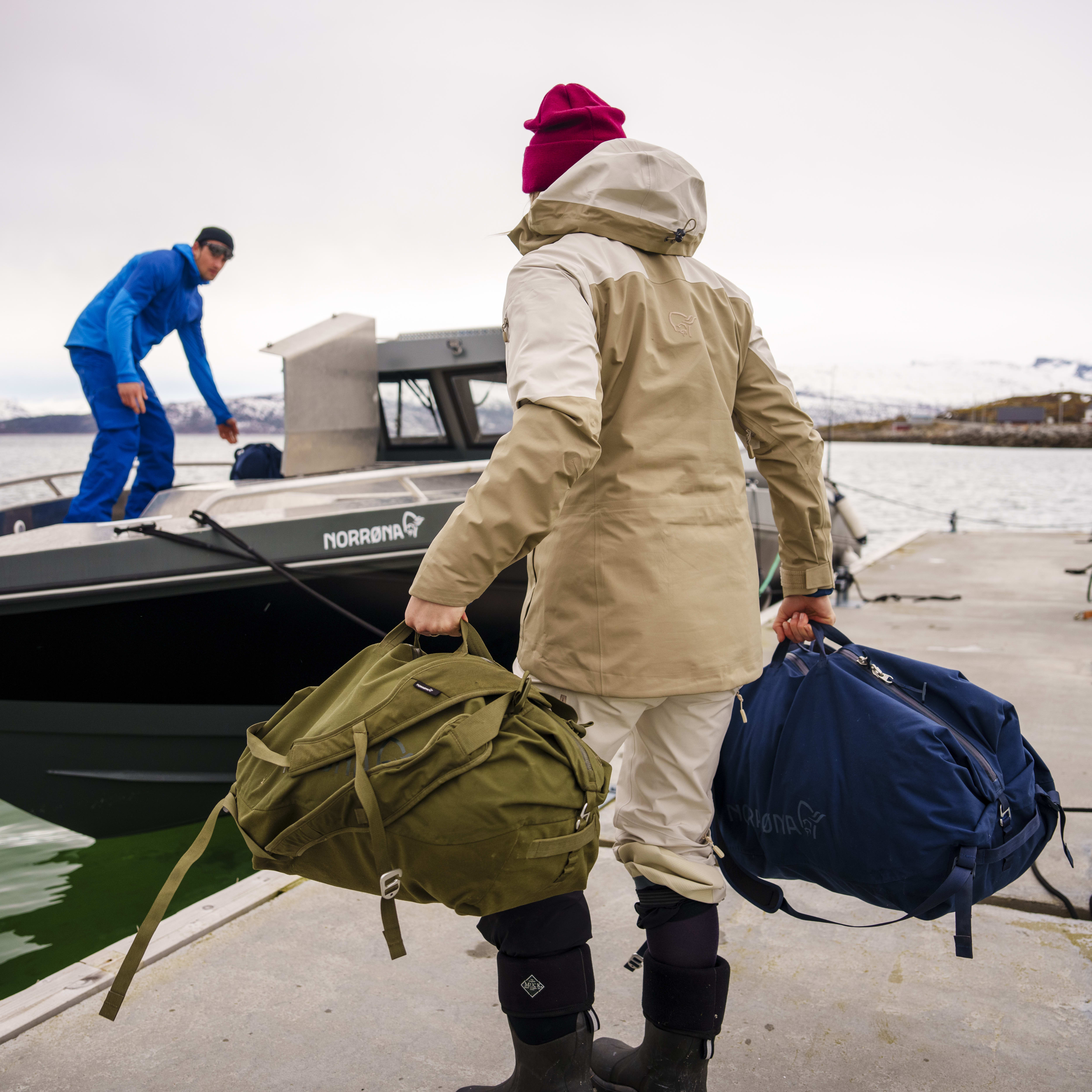A person in a Norrøna jacket and a pink hat carries two Norrøna backpacks. One backpack is green and the other is blue. They are standing on a dock with a boat behind them. The boat has the Norrøna logo on it. The person is waiting to board the boat.